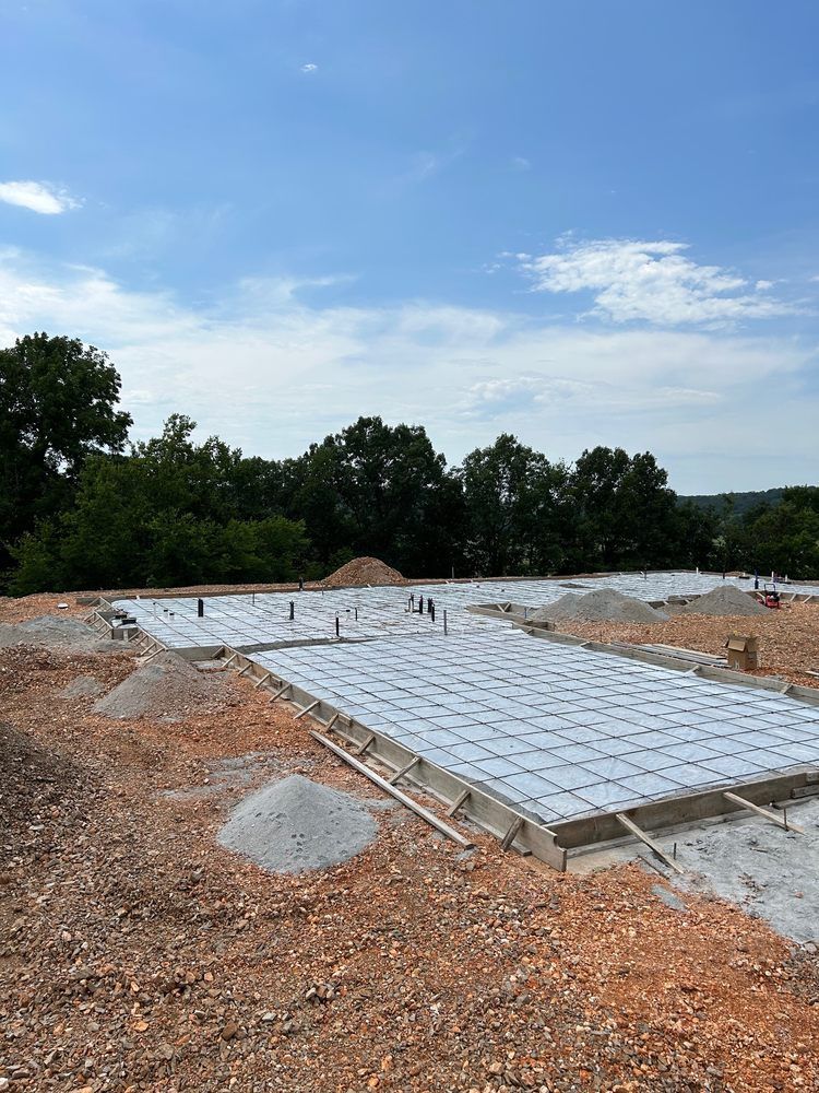 Concrete foundation slab under construction on a dirt lot with trees and blue sky in the background