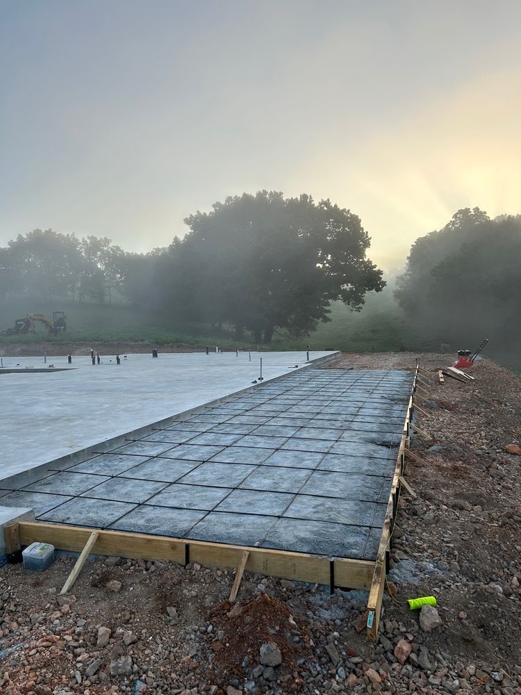 Concrete walkway under construction beside a misty pond at dawn, with trees and dirt piles nearby