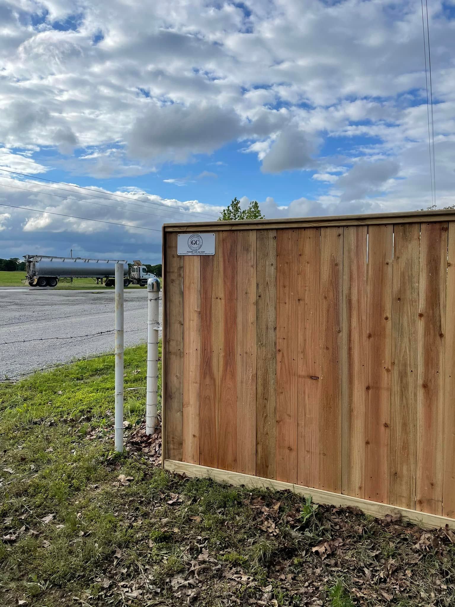 Wooden fence by a gravel road under a cloudy blue sky, with grass and a small sign on the post.