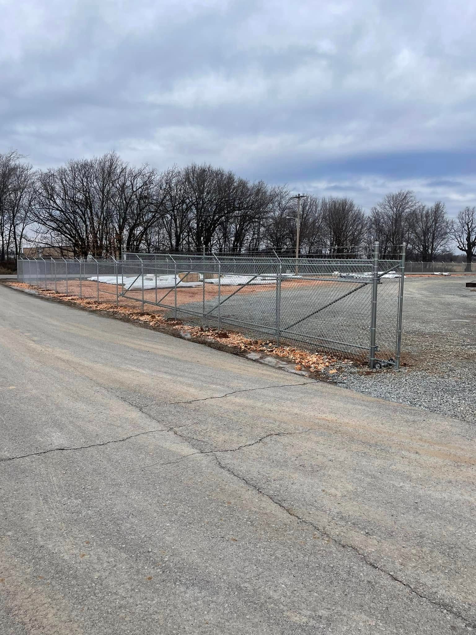 Empty gravel lot with a chain-link fence, leafless trees, and a cloudy sky