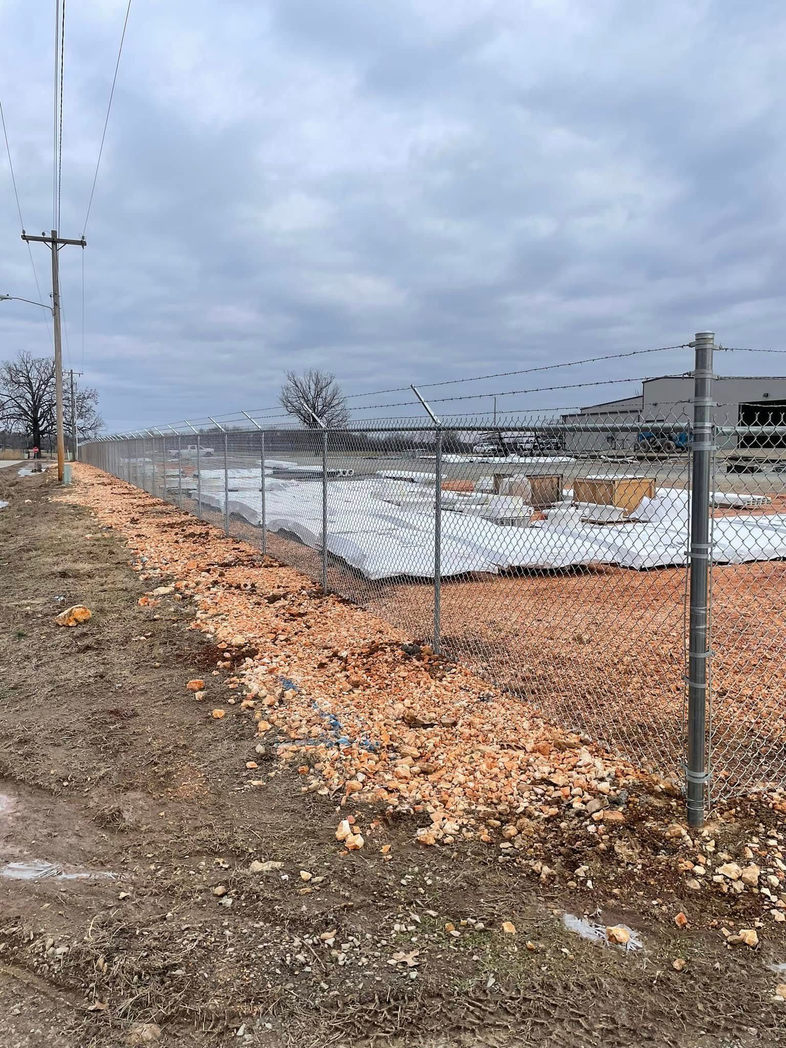 Chain-link fence around a snowy lot under a cloudy sky, with utility poles and a gravel roadside.