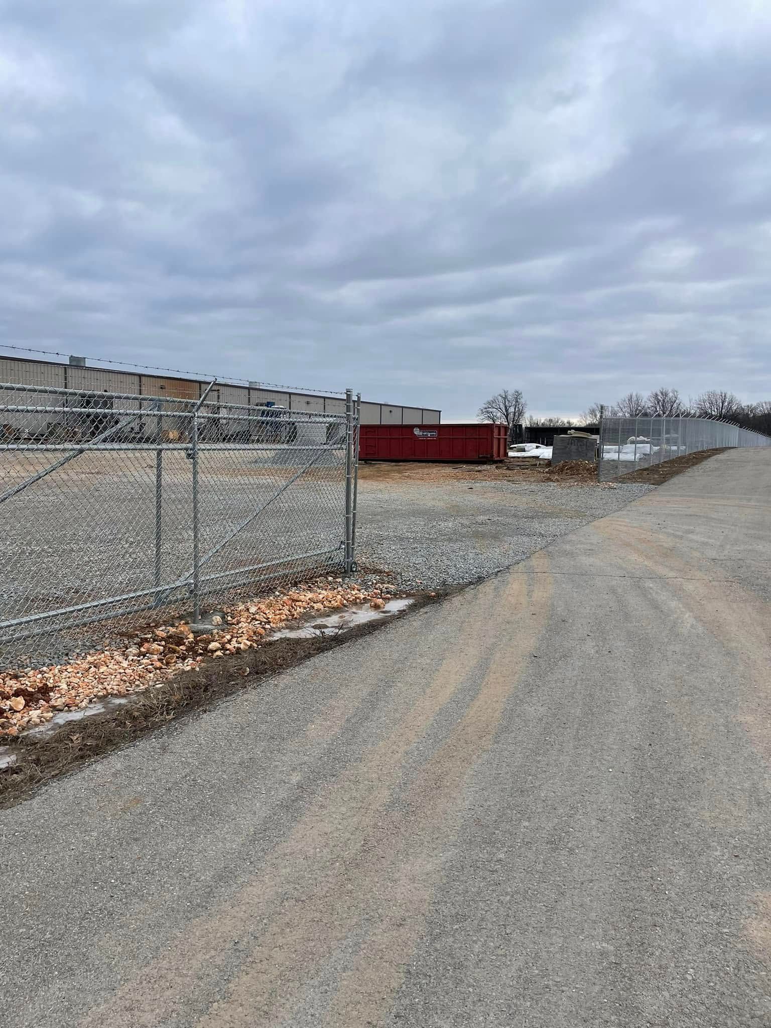Gravel road beside a chain-link fence and empty lot with a red building under cloudy skies