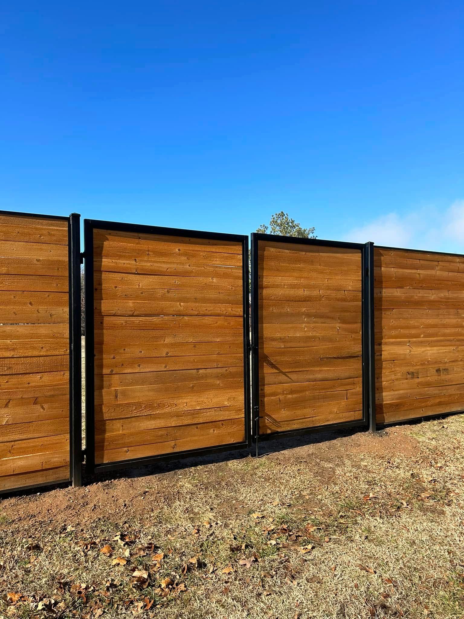 Wooden sliding gate with black metal frame along a gravel driveway under a clear blue sky