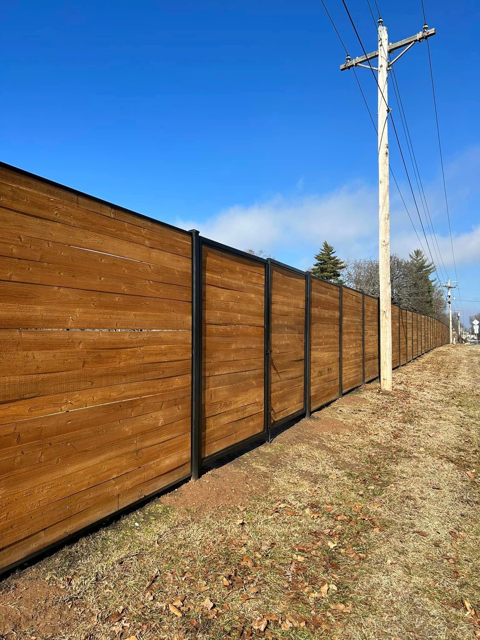 Brown wooden privacy fence beside a gravel road under a clear blue sky with utility poles