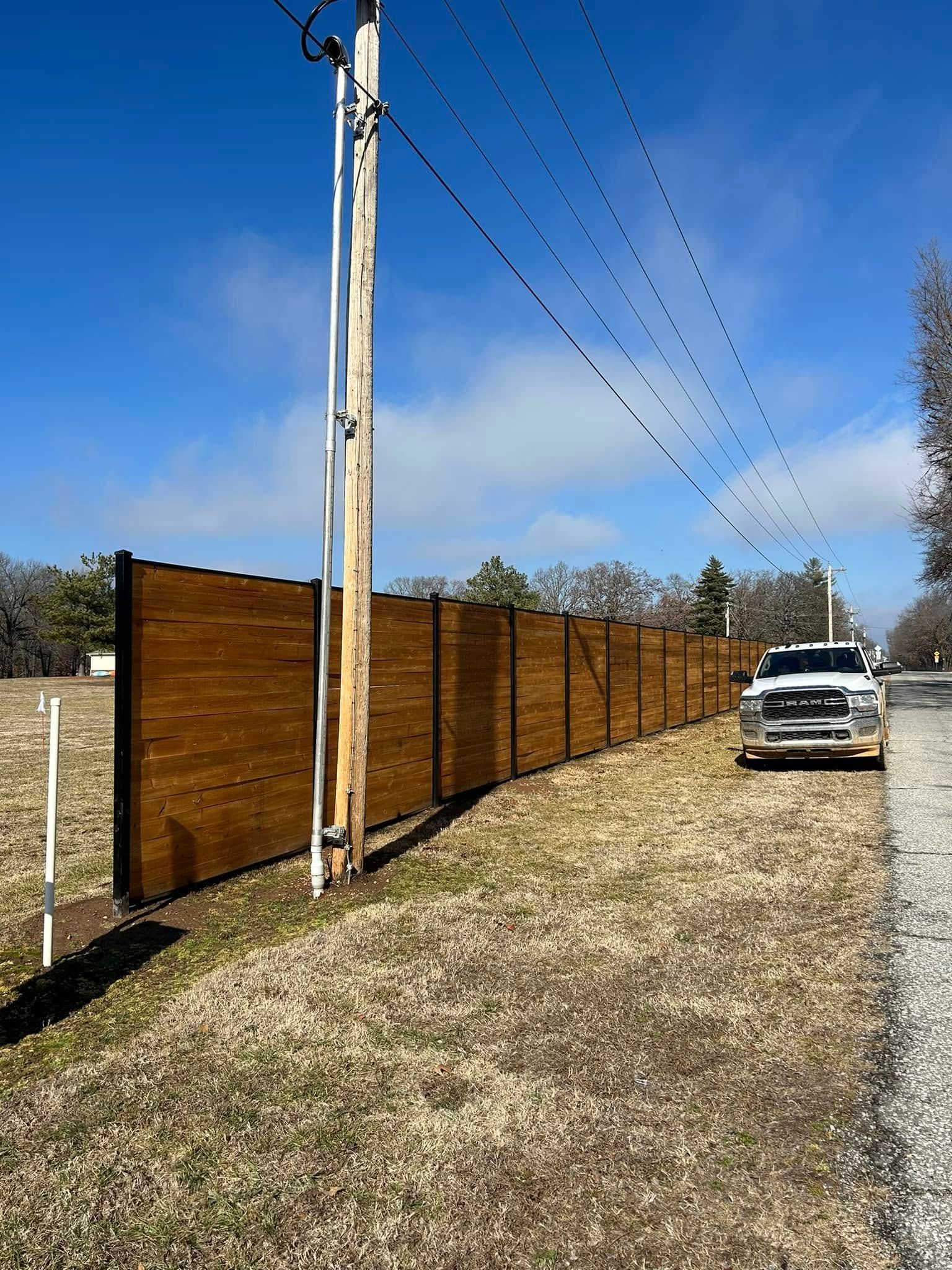 Wooden privacy fence along a gravel driveway with utility pole, parked white truck, and blue sky.