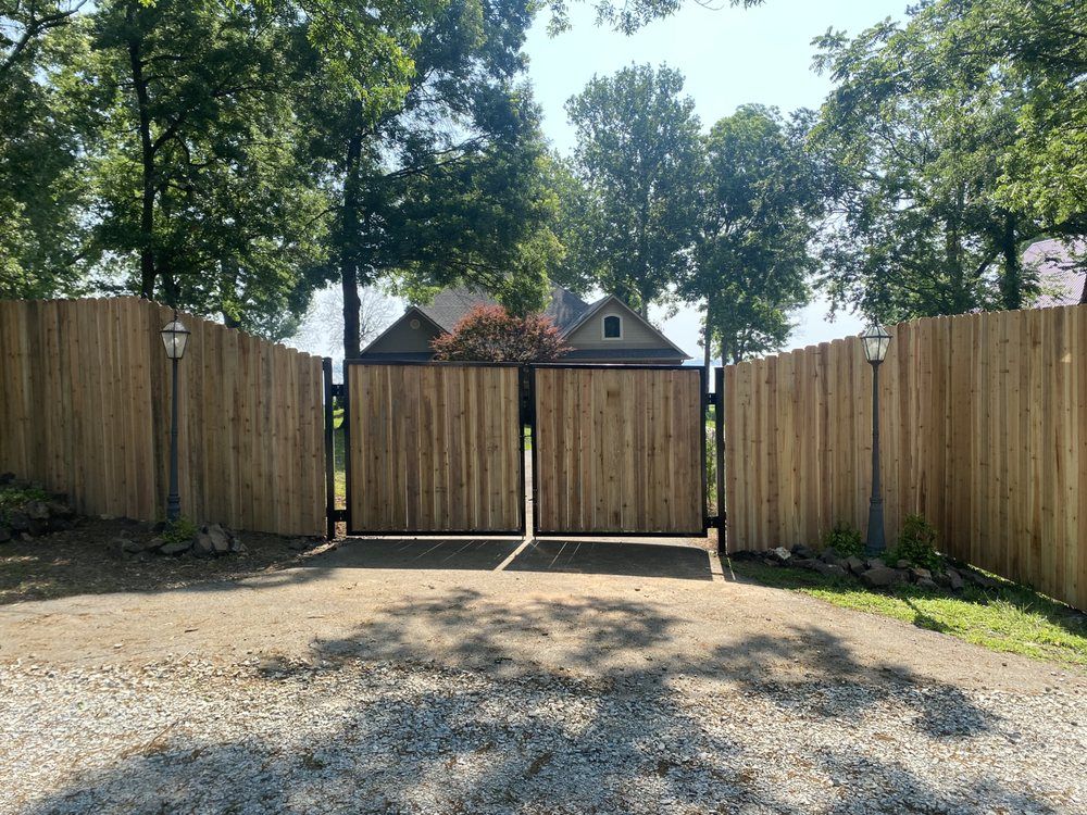 Wooden fenced driveway gate under trees, opening onto a gravel area with a house visible beyond.