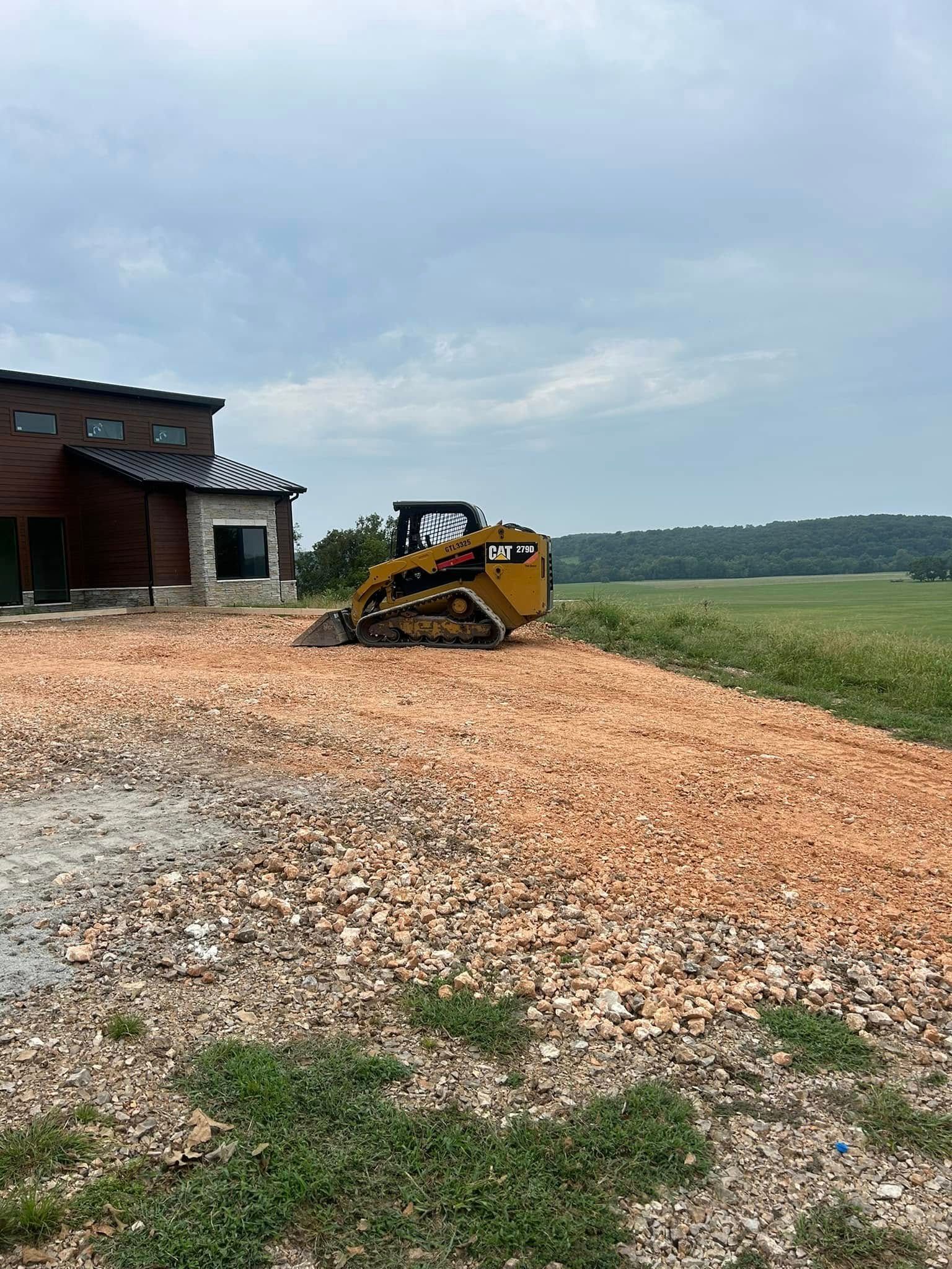 Yellow skid steer on gravel beside a barn in a rural field under a cloudy sky