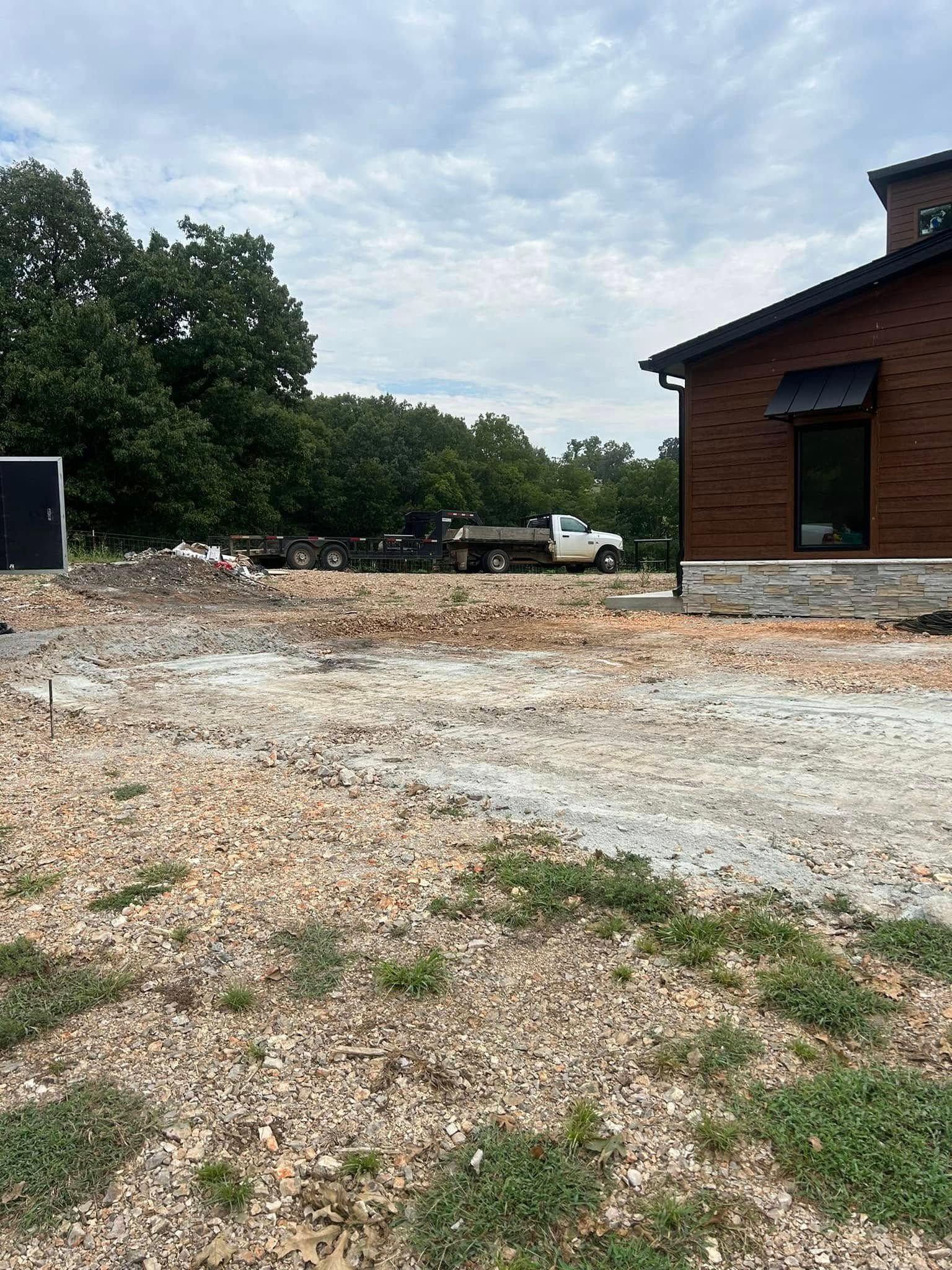 Gravel construction site beside a brick building, with trees and a cloudy sky in the background