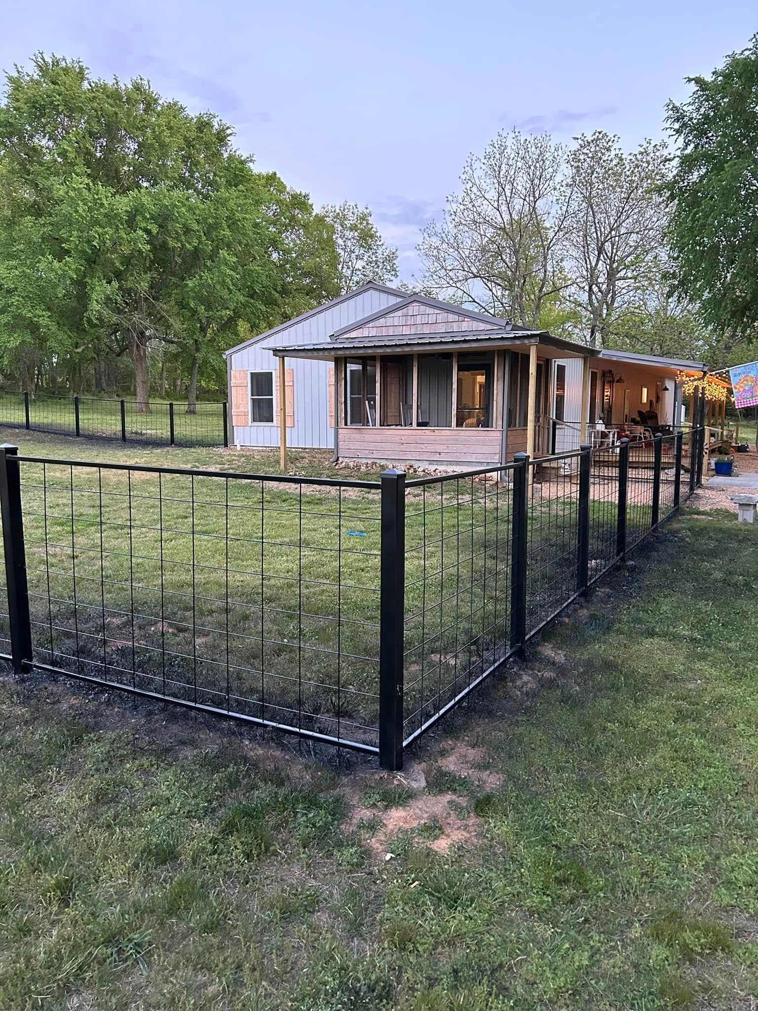 Small house with a black metal fence, surrounded by trees in a grassy yard.