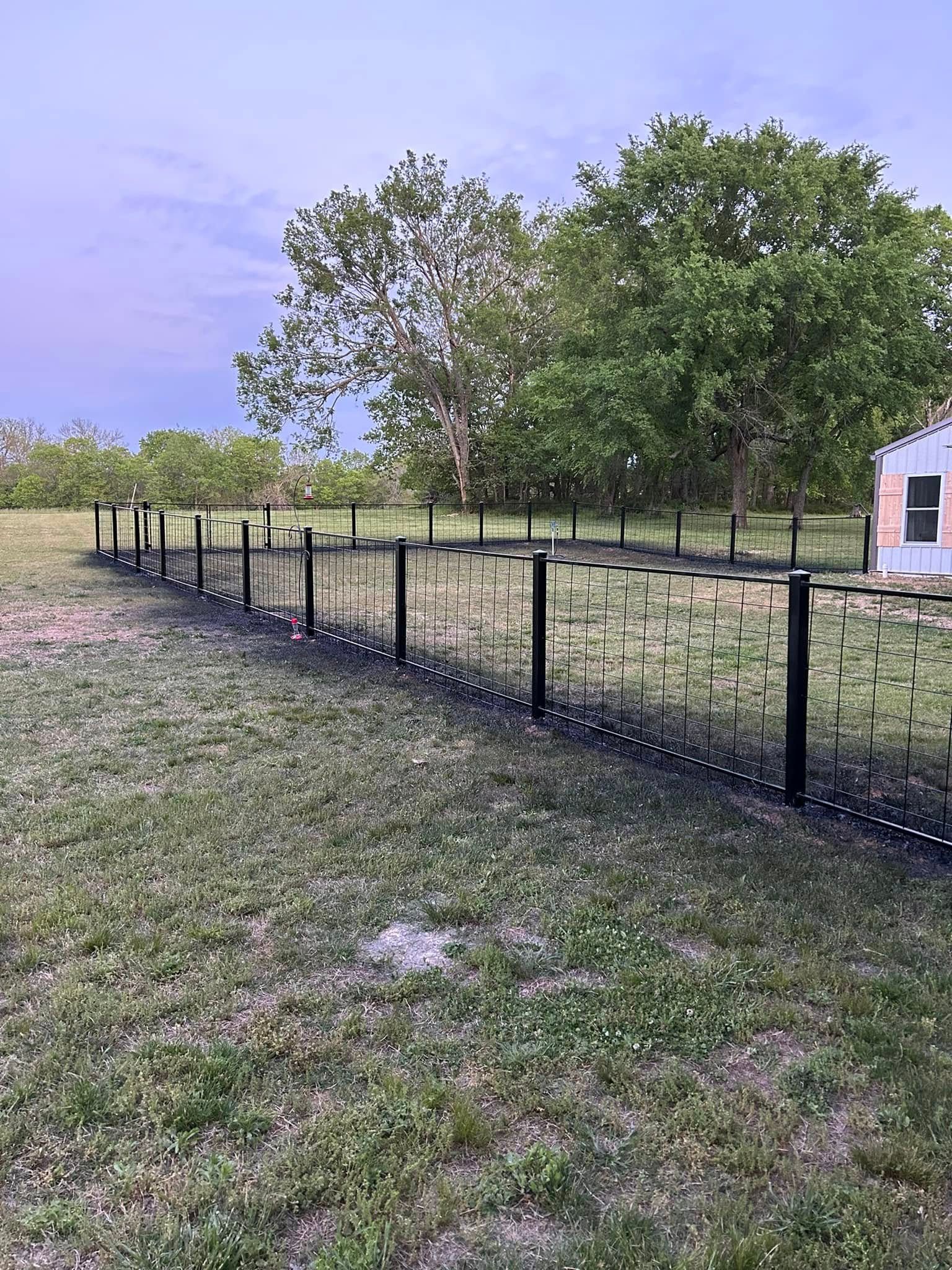 Black fence along a grassy yard with trees and a small shed under a cloudy sky