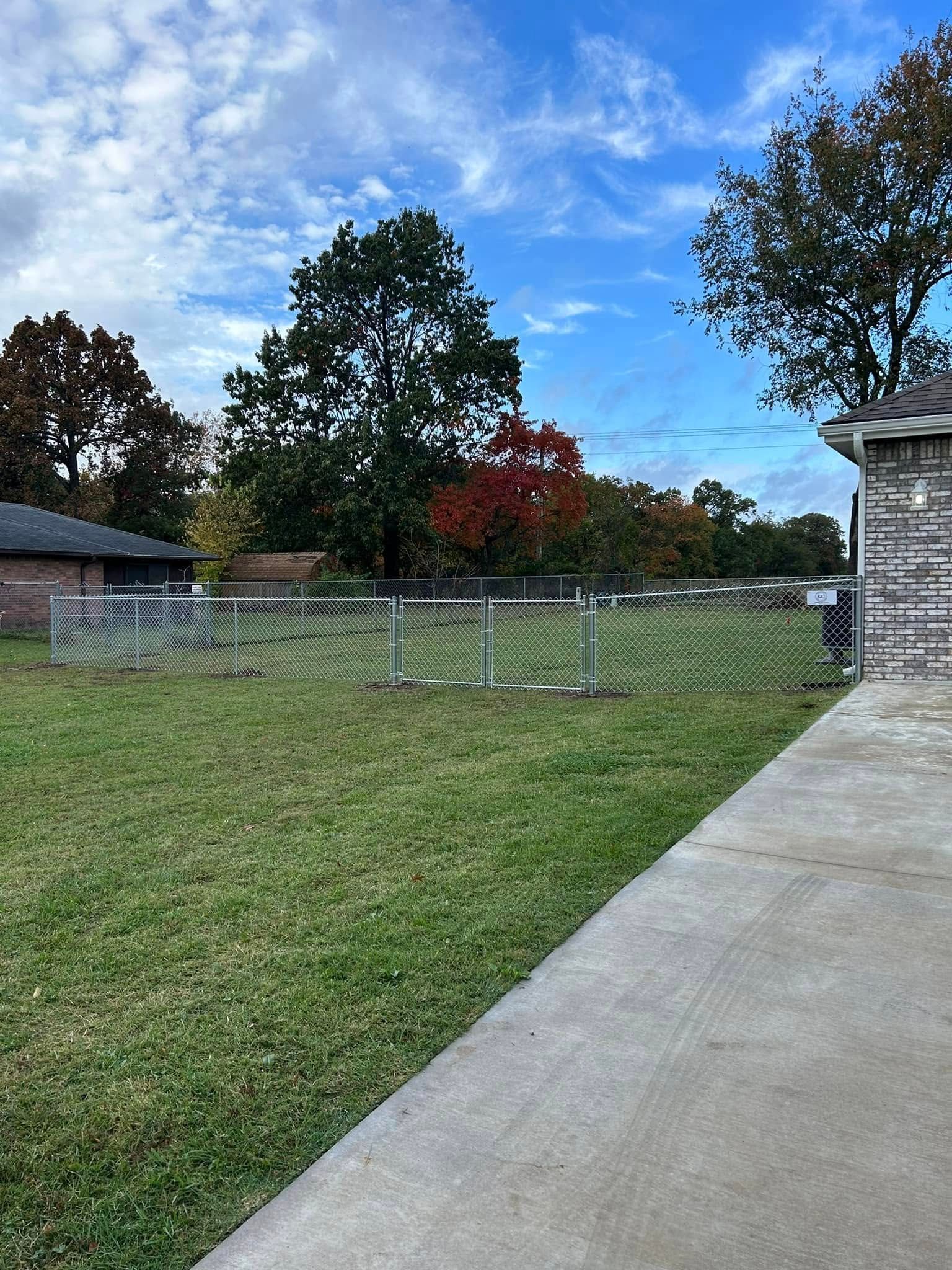 Grassy yard with a white fence, trees, and a concrete walkway beside a stone house under a blue sky.