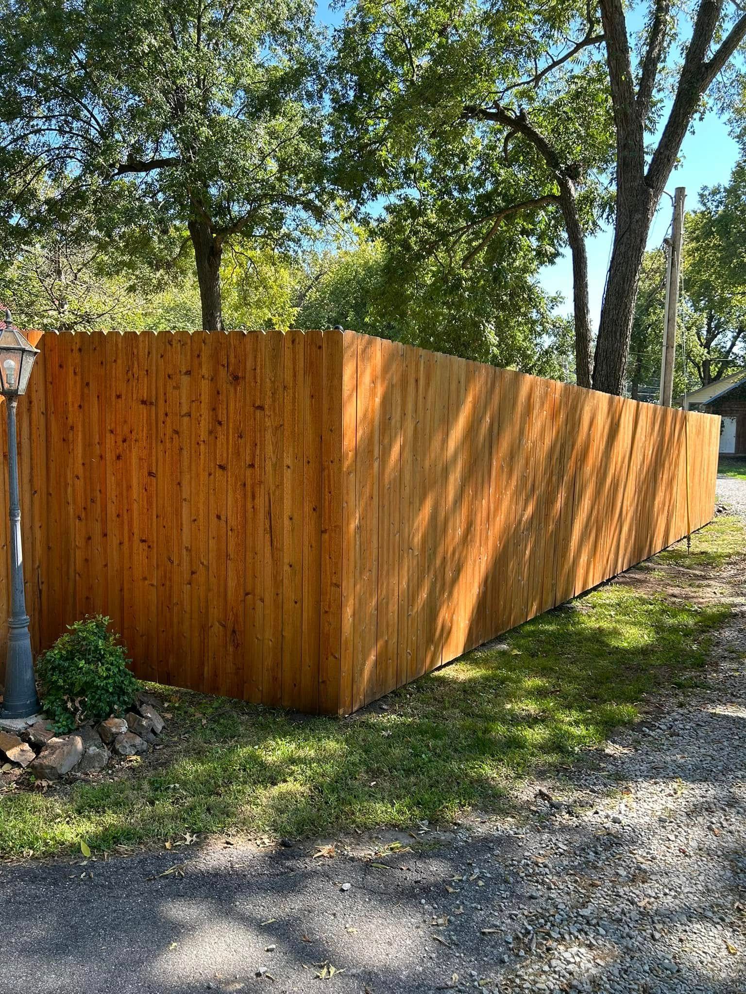 Wooden privacy fence along a gravel driveway, with trees and sunlight casting shadows.