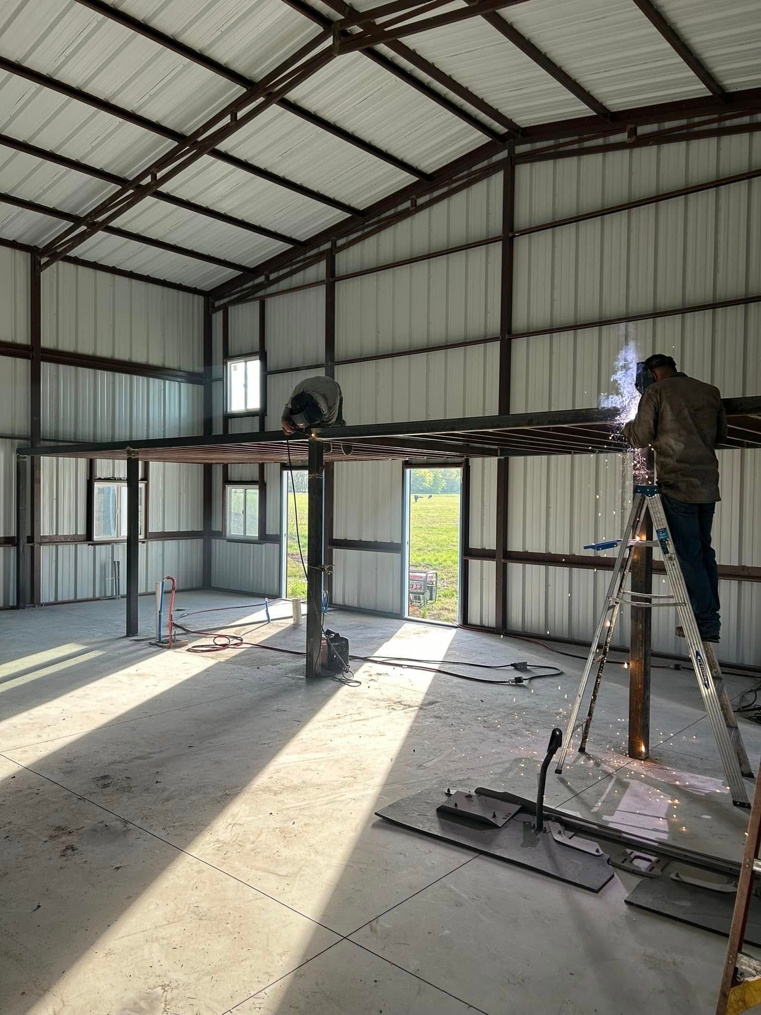 Metal workshop interior with two workers on ladders near the ceiling and open doors letting in sunlight