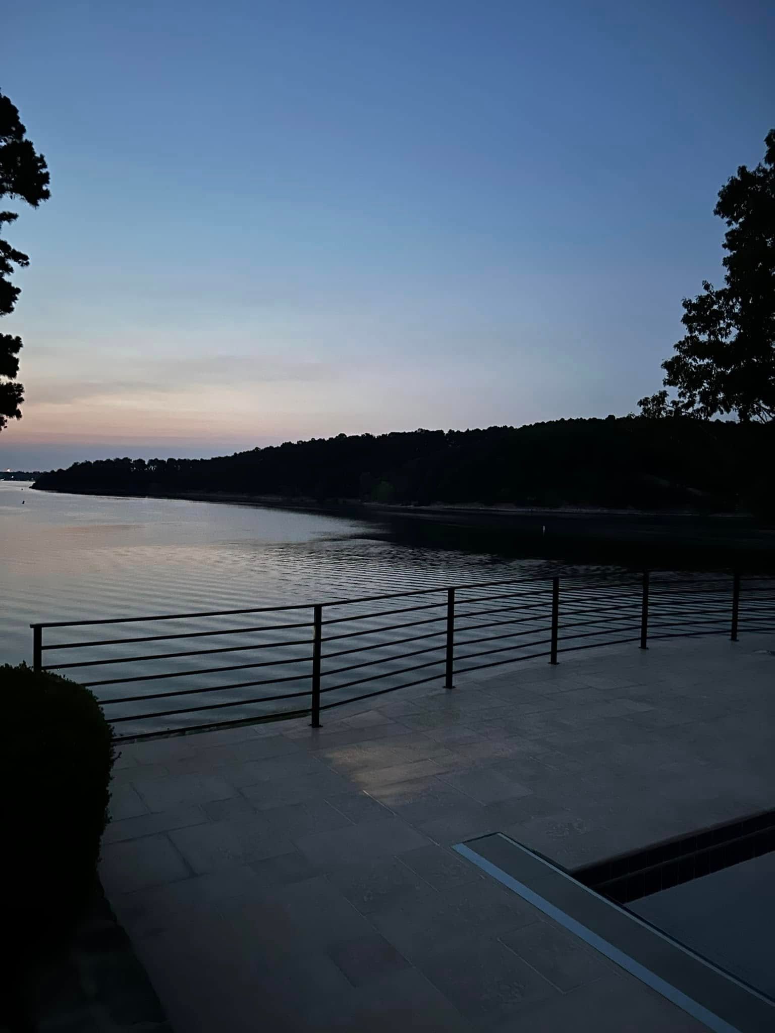 Dusk over a calm lake with silhouetted trees and a metal railing along a paved waterfront terrace.