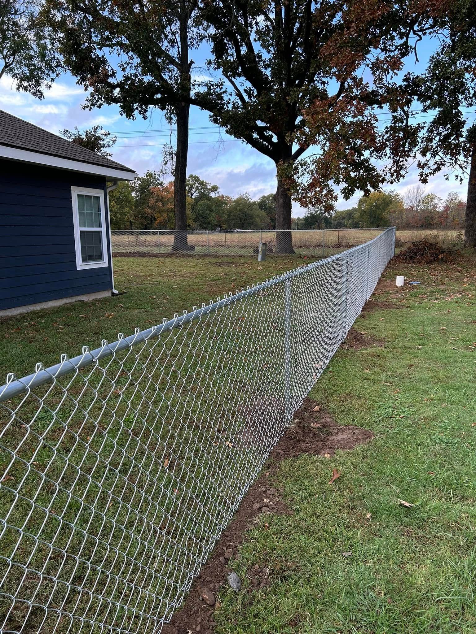 Chain-link fence beside blue house in grassy yard, stretching toward trees under a cloudy sky