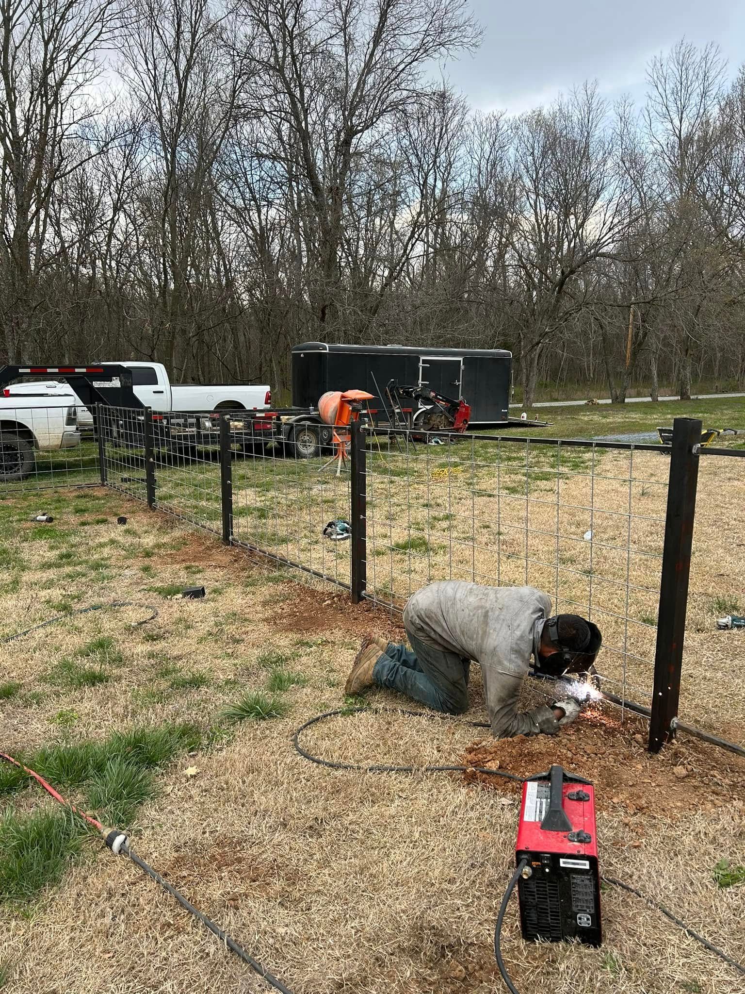 Worker kneeling by a fenced plot, using tools beside trucks and a trailer in a leafless field.