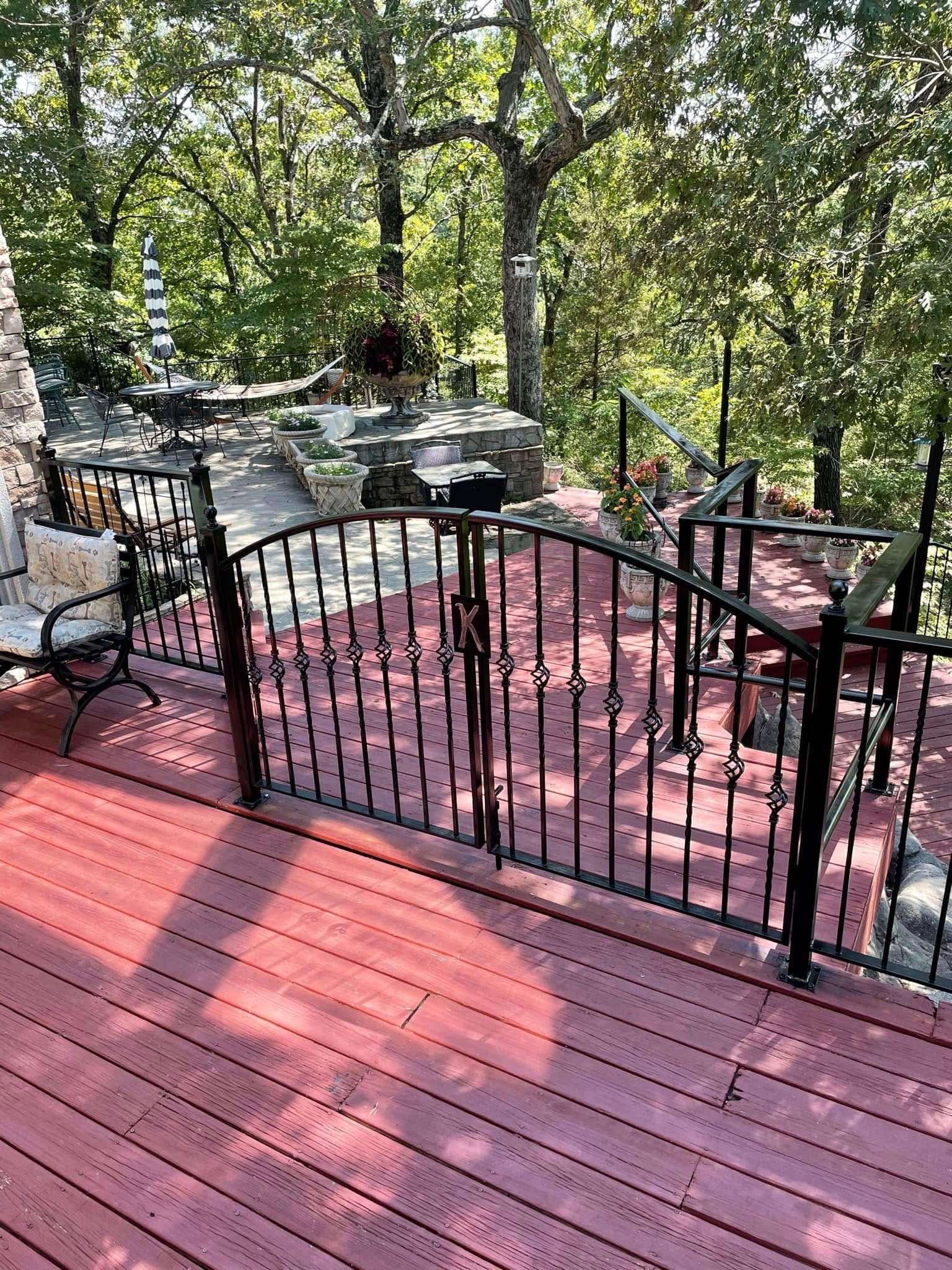 Red wooden deck with black railing, patio chairs, and stone steps beside a wooded yard.