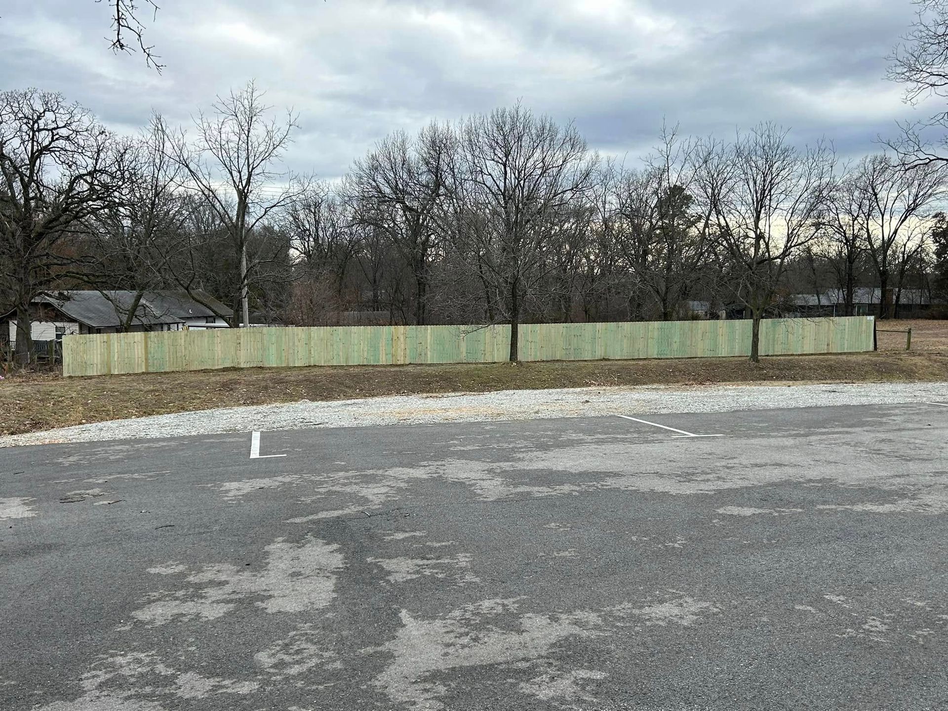Empty paved lot with a grassy edge, leafless trees, and a cloudy sky.