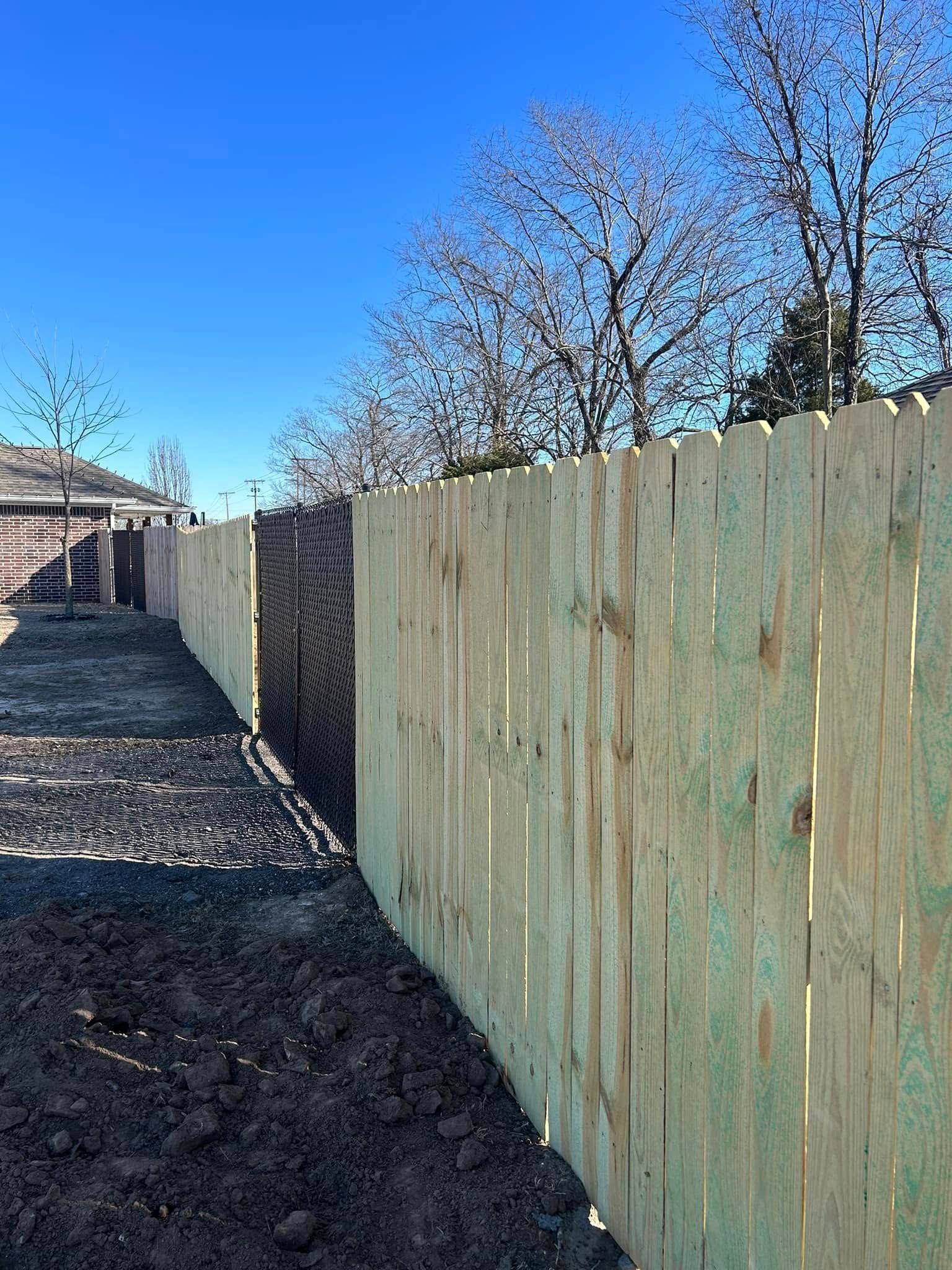 Sunlit wooden fence along a gravel path beside bare trees under a clear blue sky