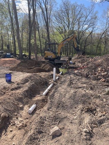 Excavator at a wooded construction site beside a trench with pipes and dirt piles
