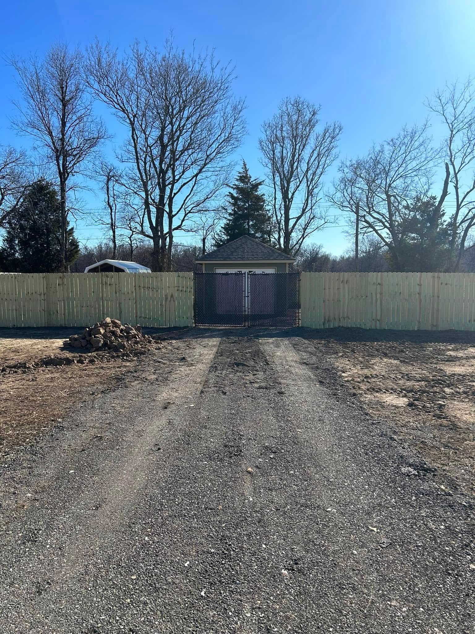 Gravel driveway leading to a closed wooden gate and fence under a clear blue sky