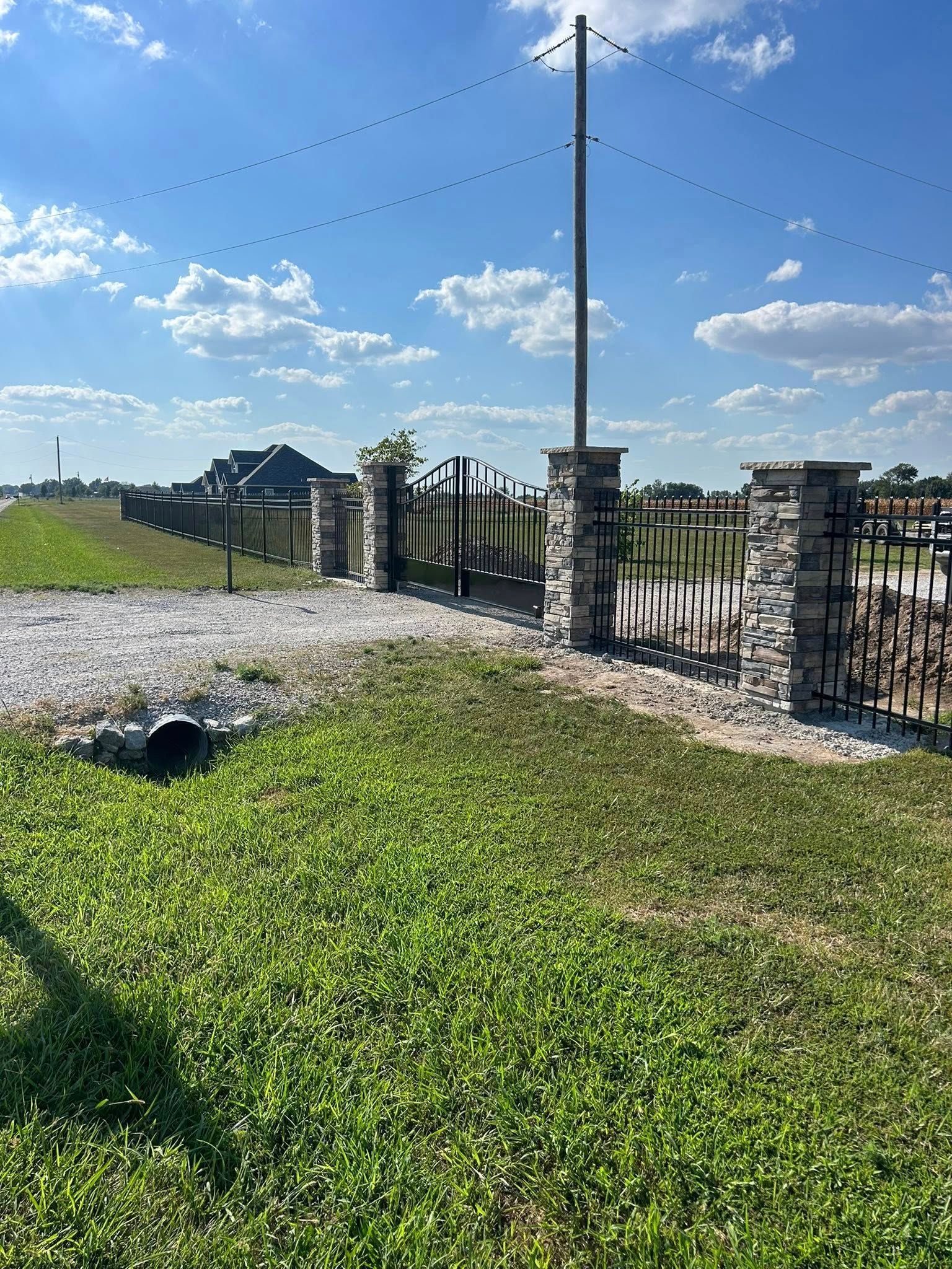 Rural gate with stone pillars, gravel driveway, utility pole, and open grassy field under a blue sky