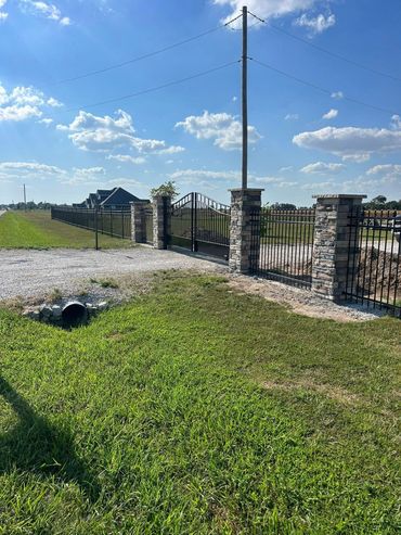 Rural gate with stone pillars, gravel driveway, utility pole, and open grassy field under a blue sky