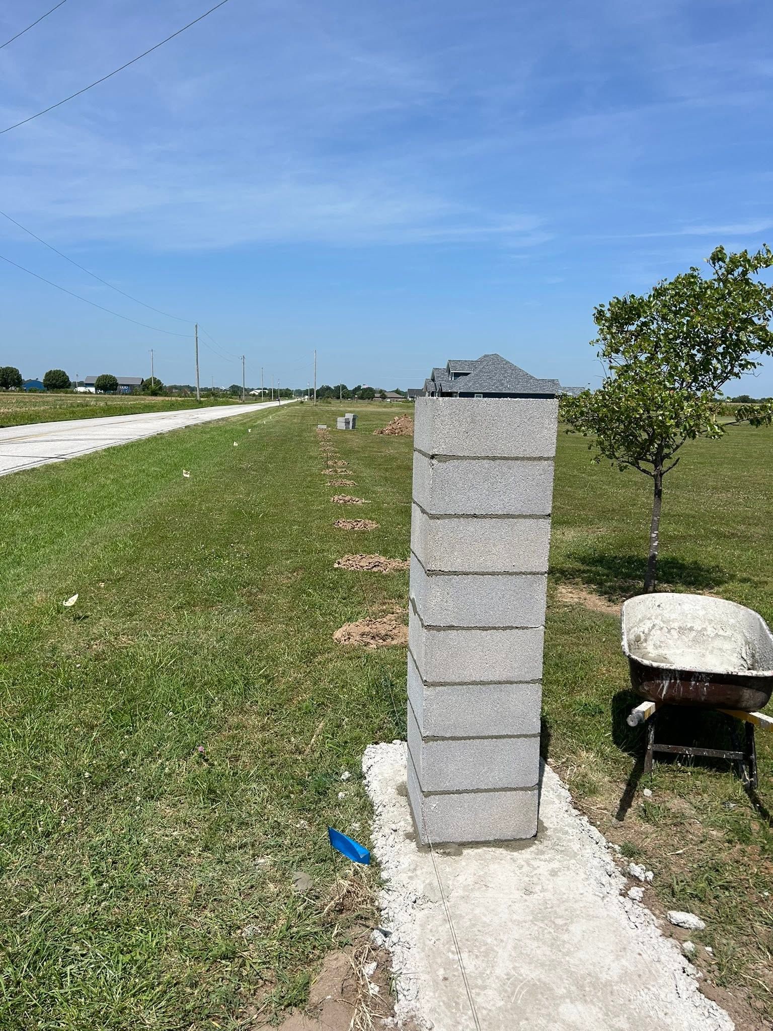Concrete pillar beside a grassy roadside under a clear blue sky, with a wheelbarrow nearby.