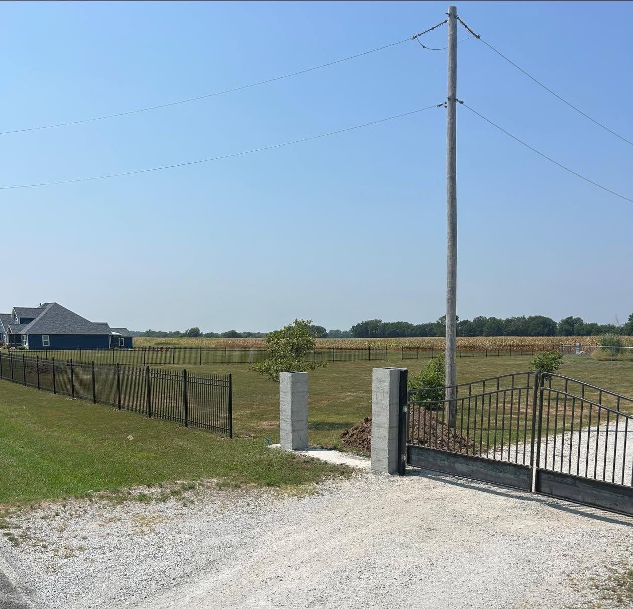 Rural driveway gate beside a gravel road, fenced field, and utility pole under a clear blue sky
