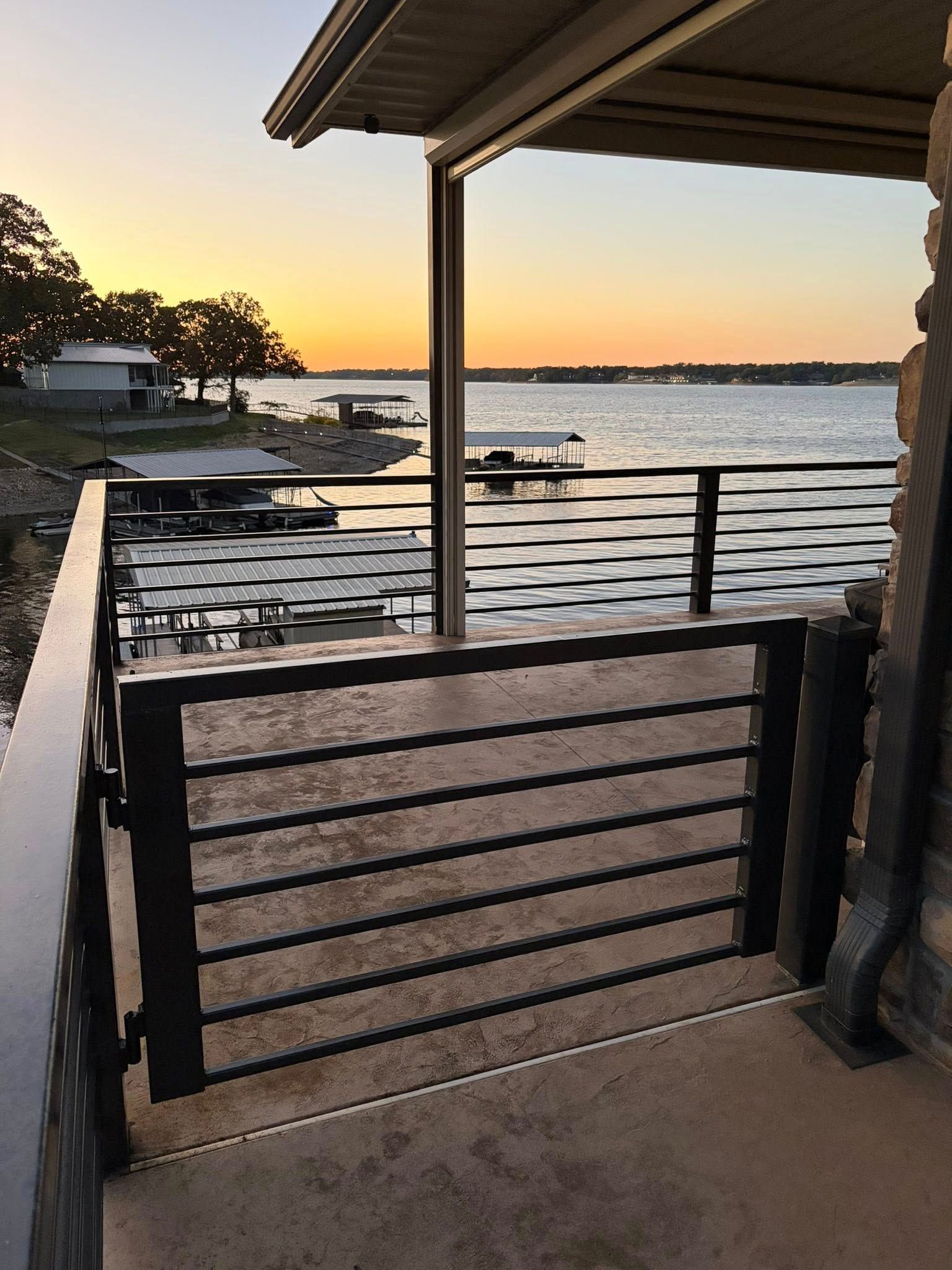 Sunset view from a covered dock over calm water, with a railing and boat in the distance