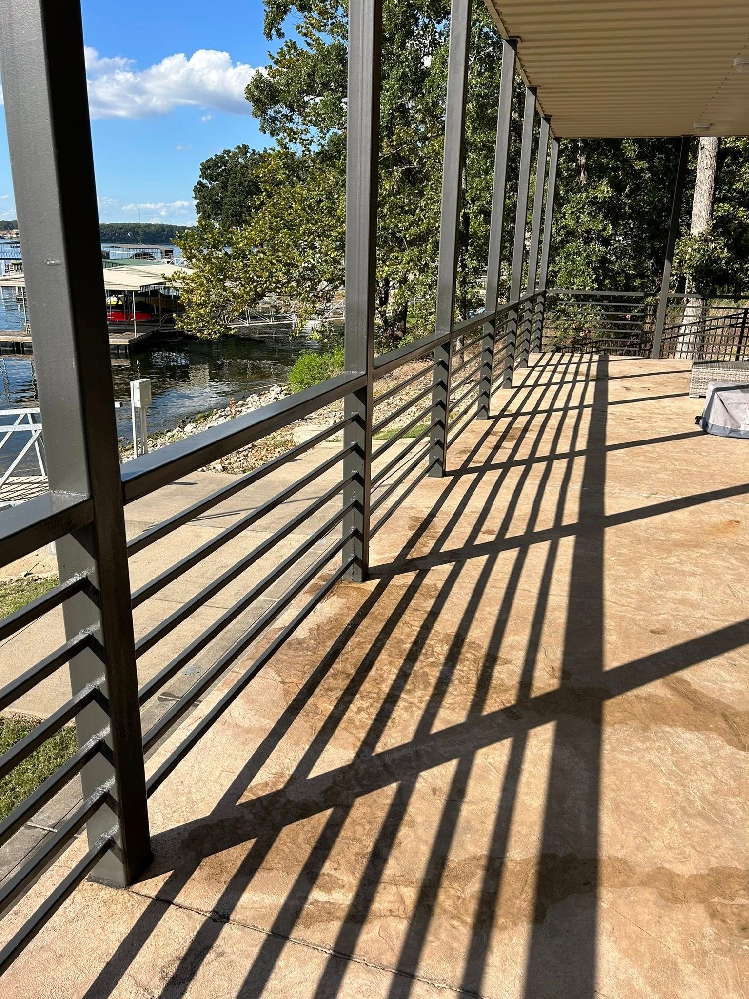 Sunlit balcony walkway with black railings, casting long shadows toward a tree-lined waterfront view.