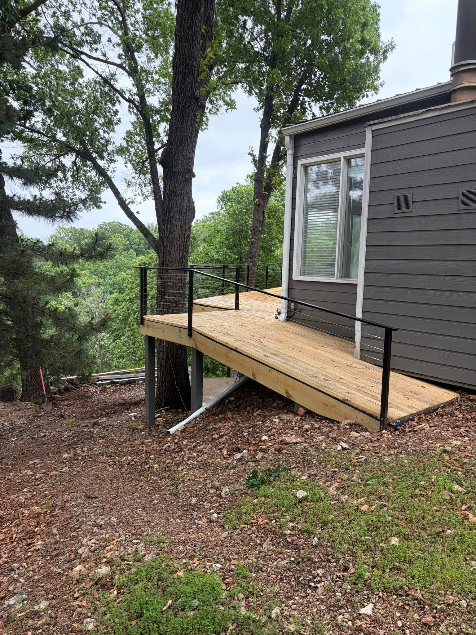 Wooden ramp with black railings leading to a gray house on a wooded hillside