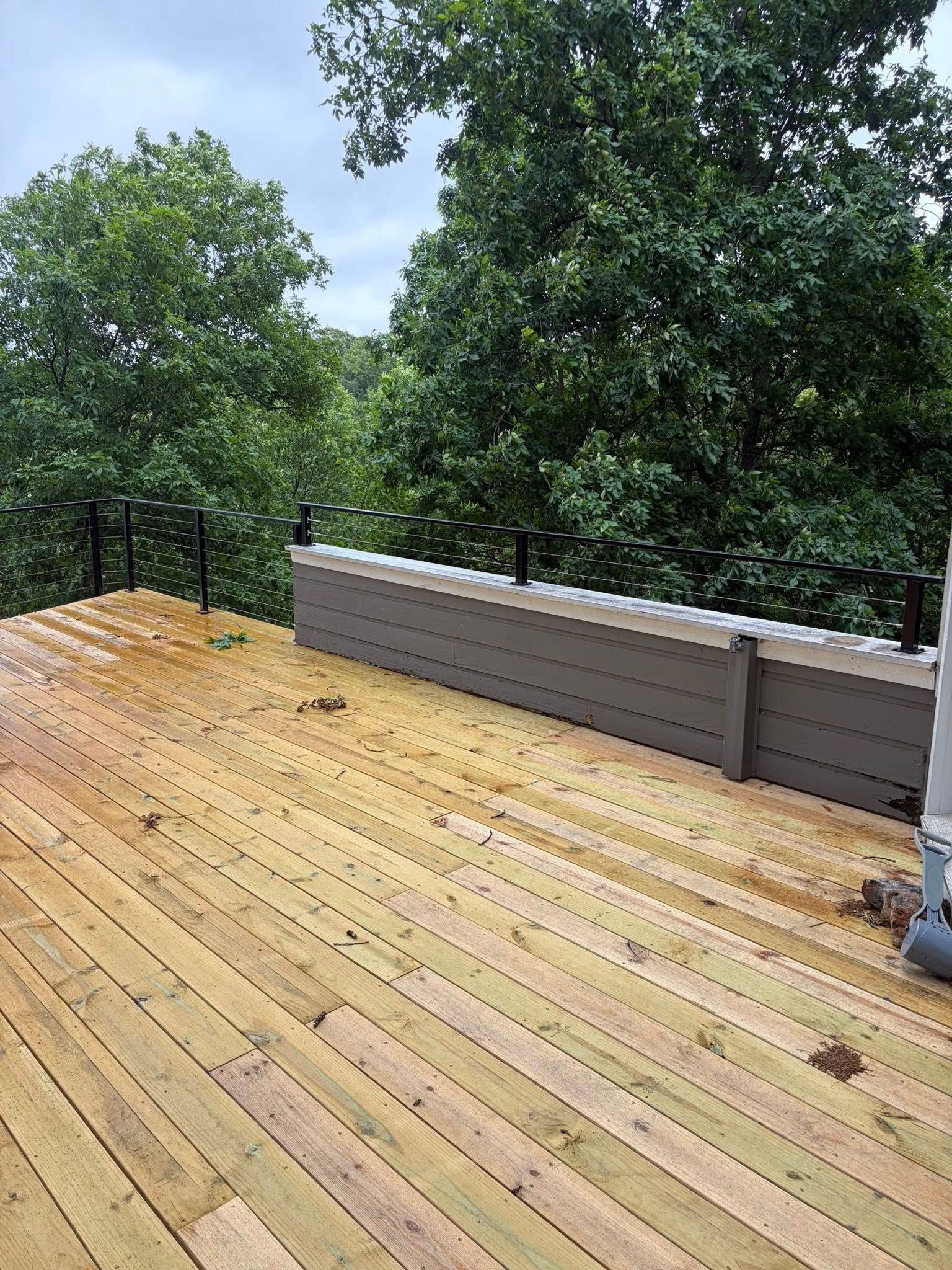 Wooden deck with a gray railing, surrounded by green trees under a bright sky