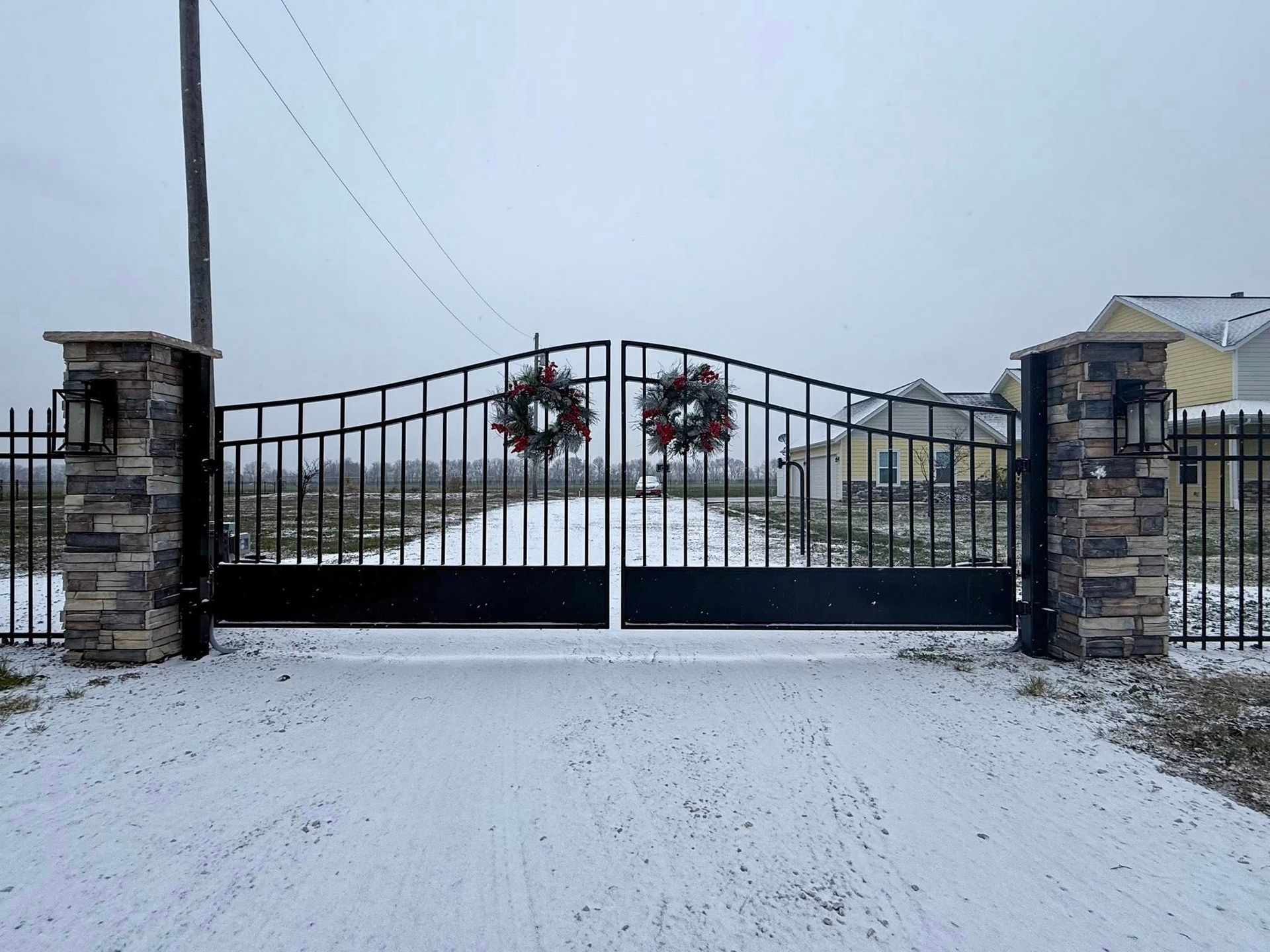 Snow-covered driveway gate with stone pillars and holiday wreaths on a cloudy day