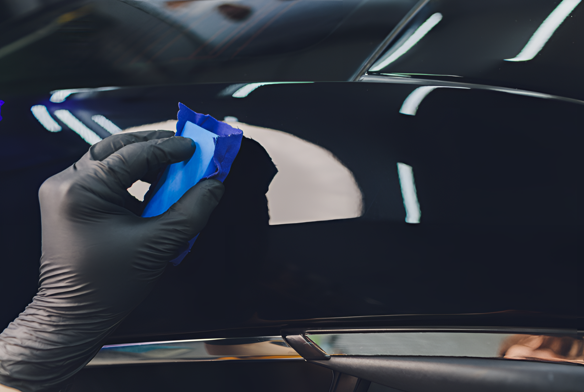 A gloved hand uses a blue applicator sponge to apply a protective coating to a shiny black car panel.