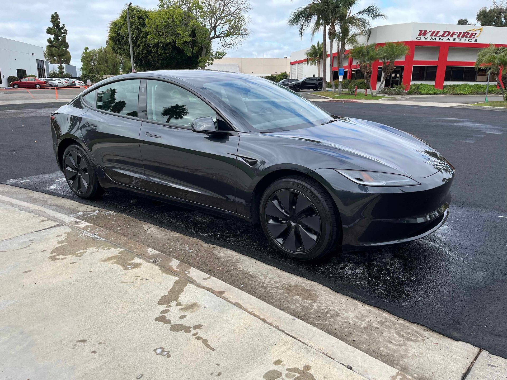A dark gray Tesla Model 3 parked on the side of a street near a building.