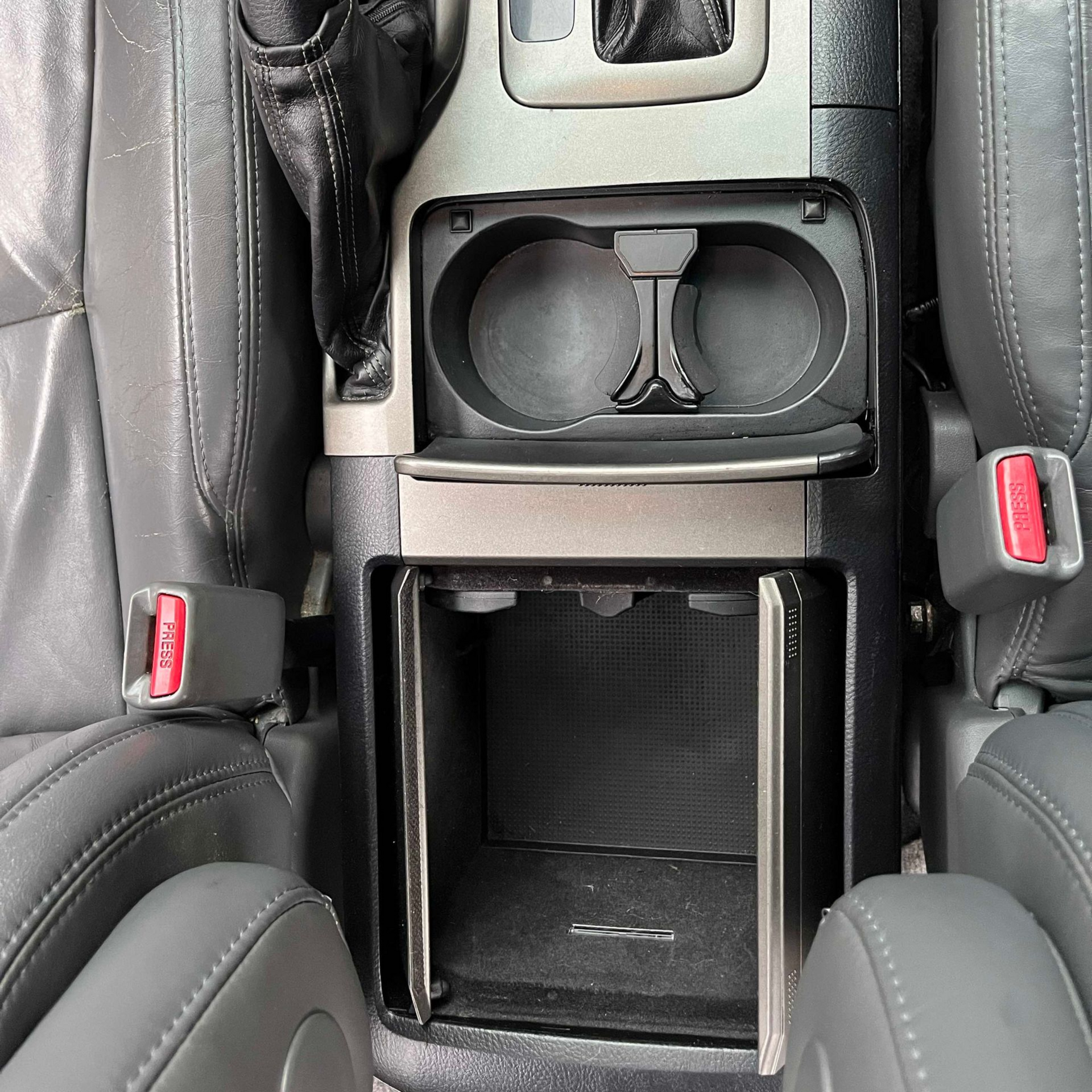 Car interior center console with dual cup holders and an open storage compartment, flanked by black seats.