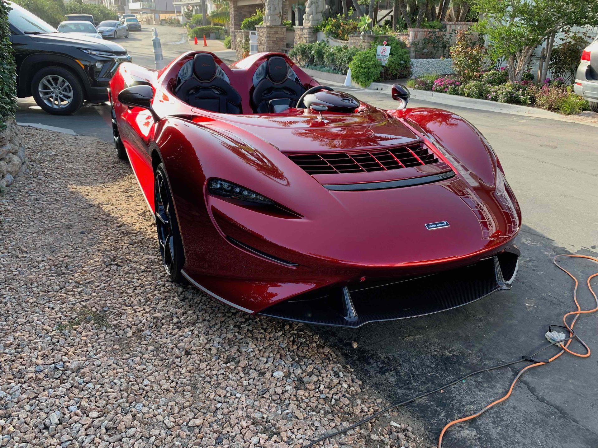 A deep red McLaren Elva roadster parked on a gravel shoulder near a residential street.