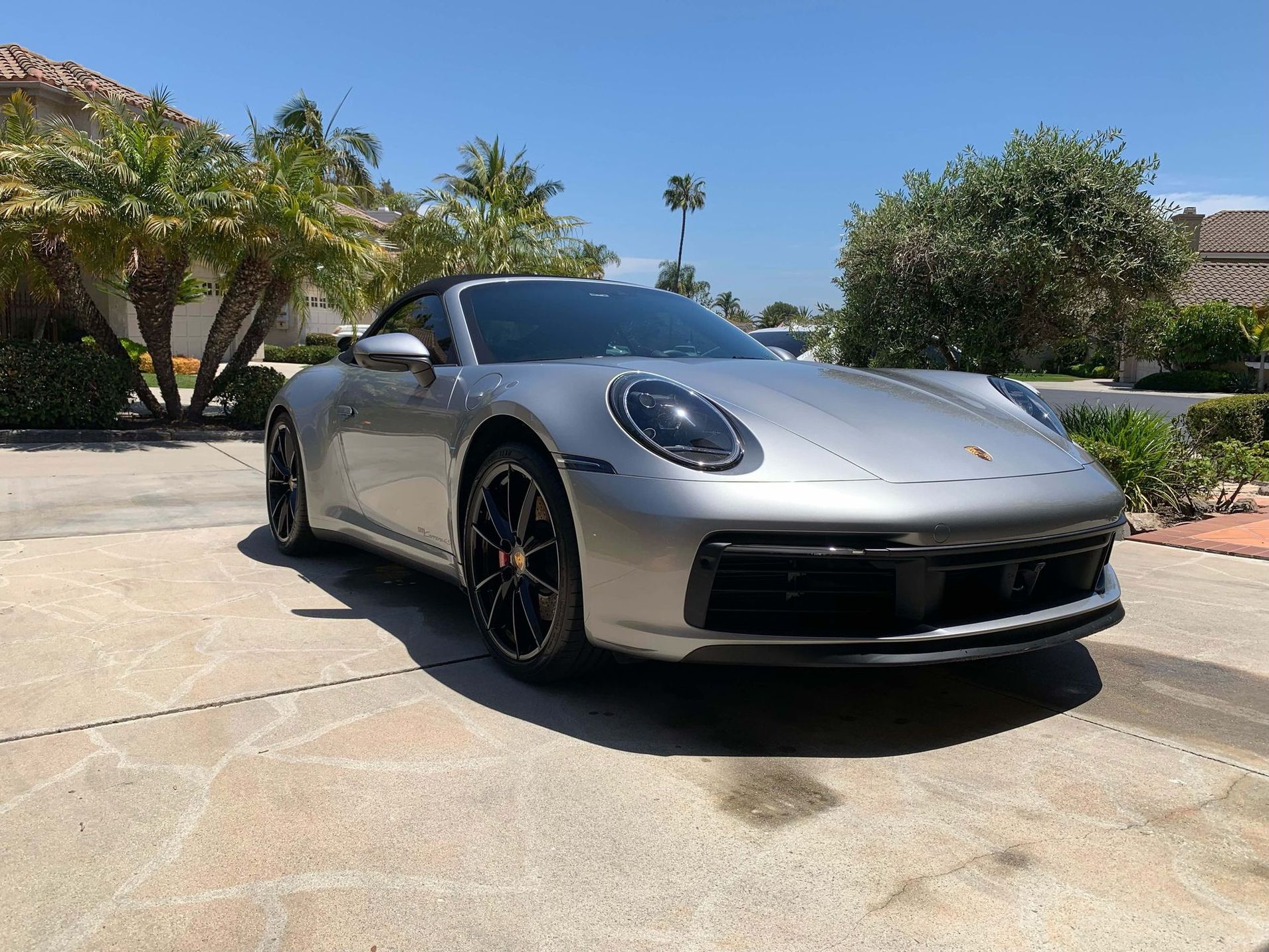 A silver Porsche 911 convertible parked on a sunny residential concrete driveway with palm trees in the background.