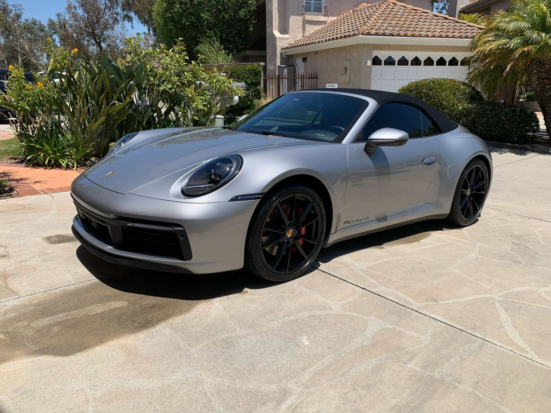 A silver Porsche 911 convertible with black wheels parked on a sunny concrete driveway in front of a house.