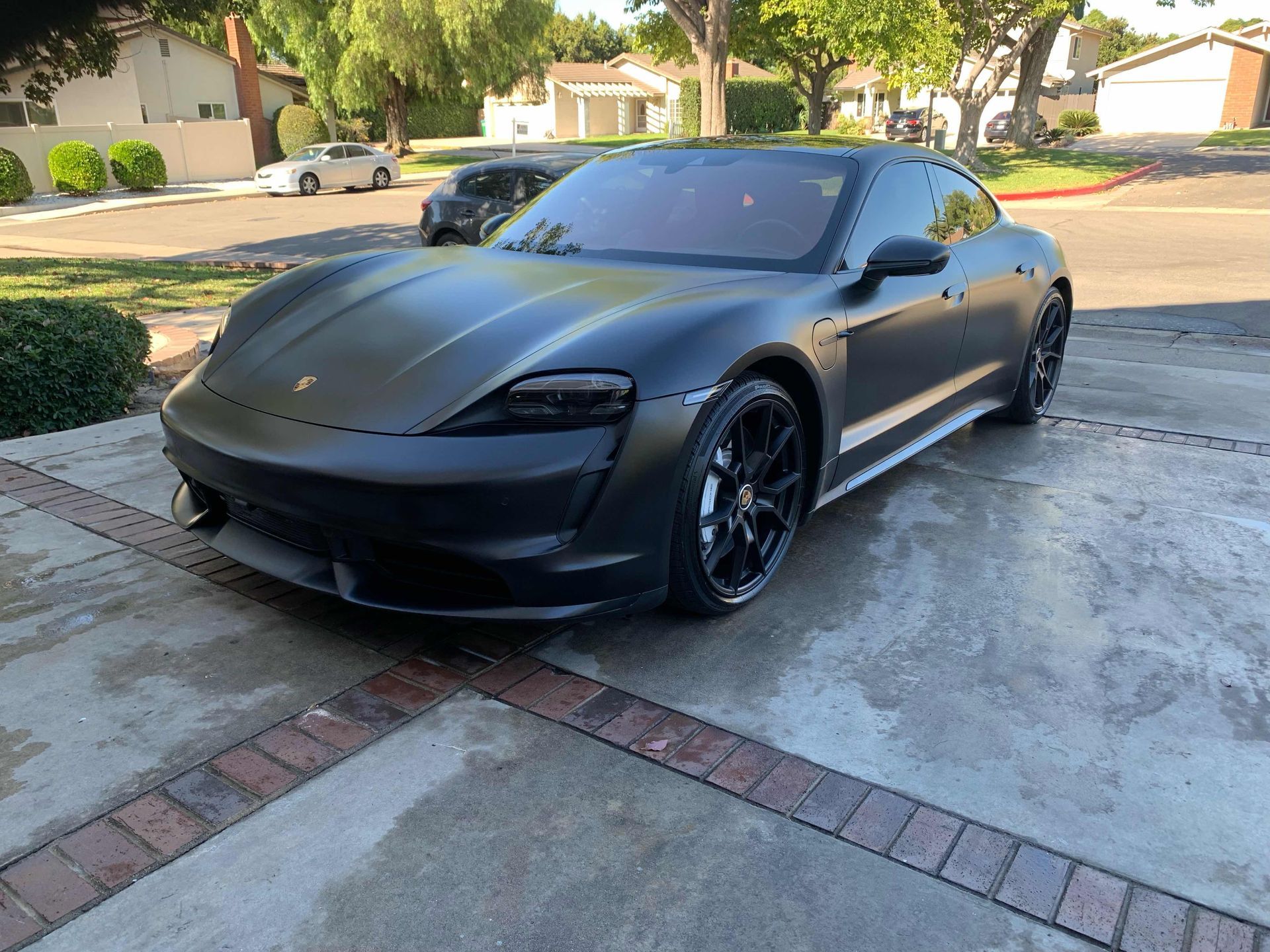 A matte black Porsche Taycan parked on a concrete driveway in a suburban neighborhood.