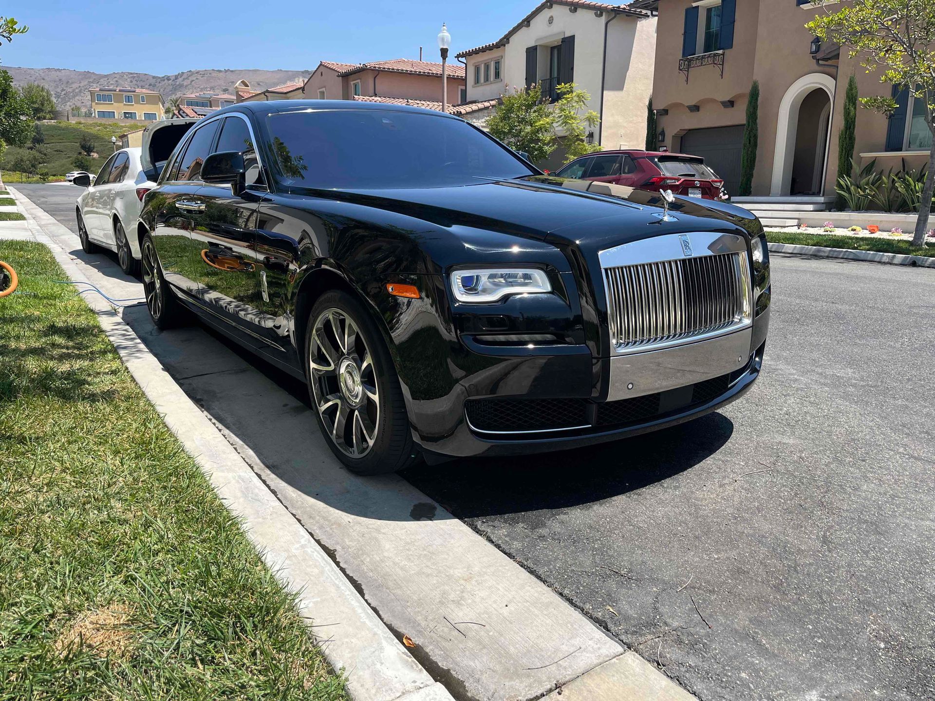 A sleek, black Rolls-Royce sedan parked on a sunny suburban street next to a curb and green lawn.