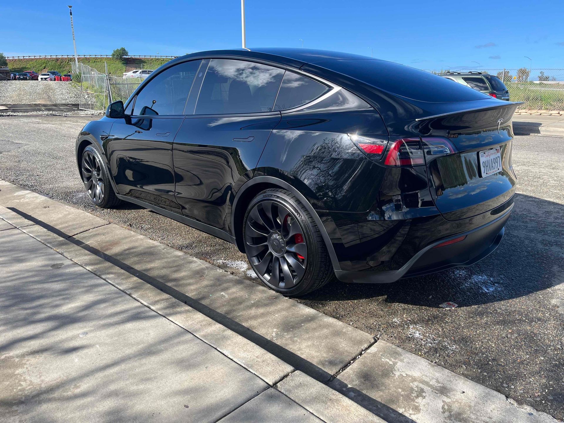 A black Tesla Model Y parked on a gravel lot next to a concrete sidewalk.