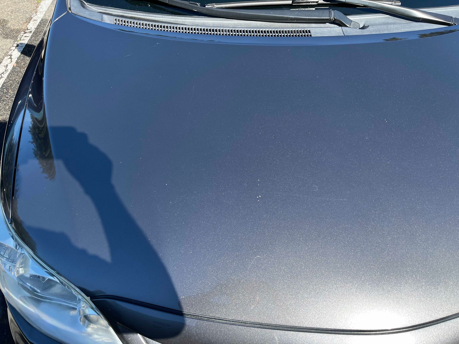 A high-angle view of a dark metallic grey car hood, showing the reflection of a person taking the photo.