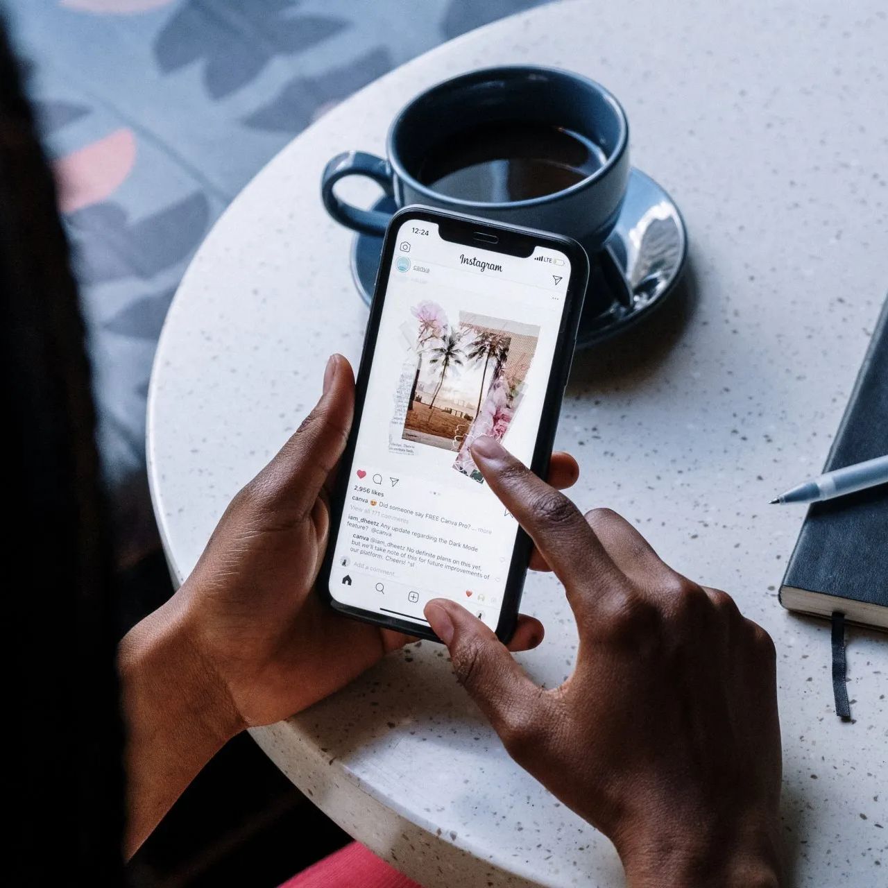 Person using a smartphone at a table with a cup of coffee and a notebook.