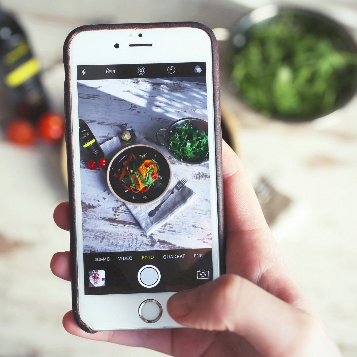 Smartphone taking photo of a bowl of food on a wooden table, tomatoes, and greens in the background.