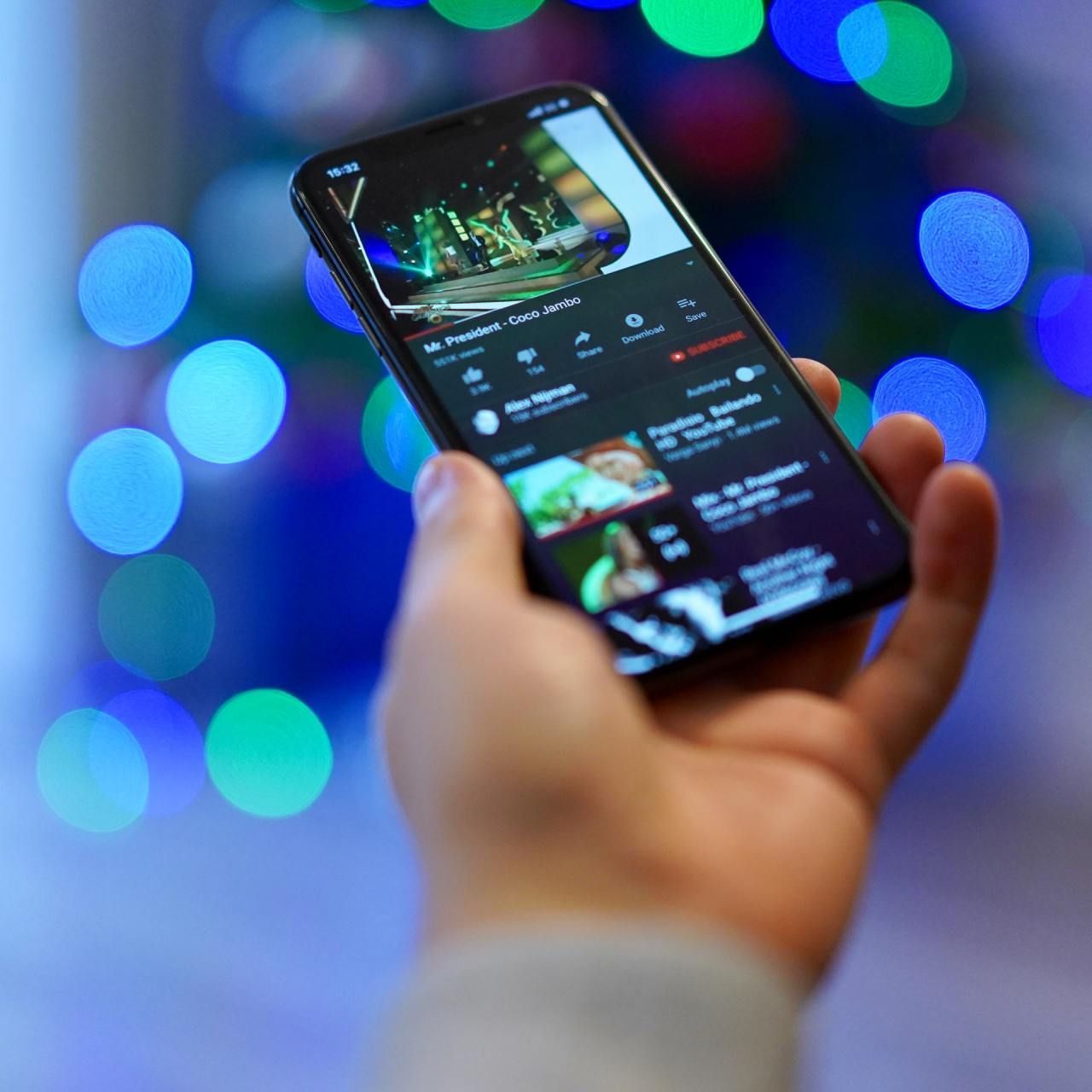 Hand holding a smartphone displaying a YouTube video with a bokeh background of blue and green lights.
