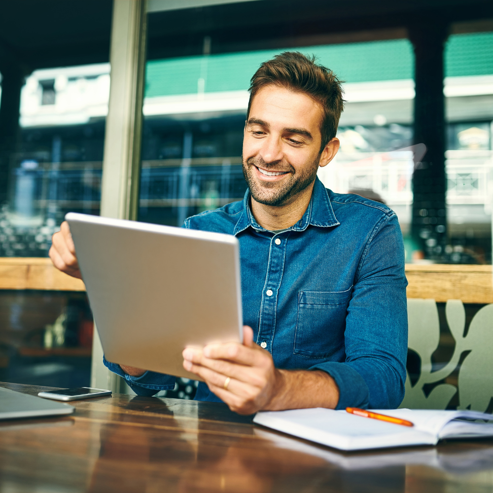 Man smiling, looking at a tablet, seated at a table near a window. Notebook and pen in front of him.