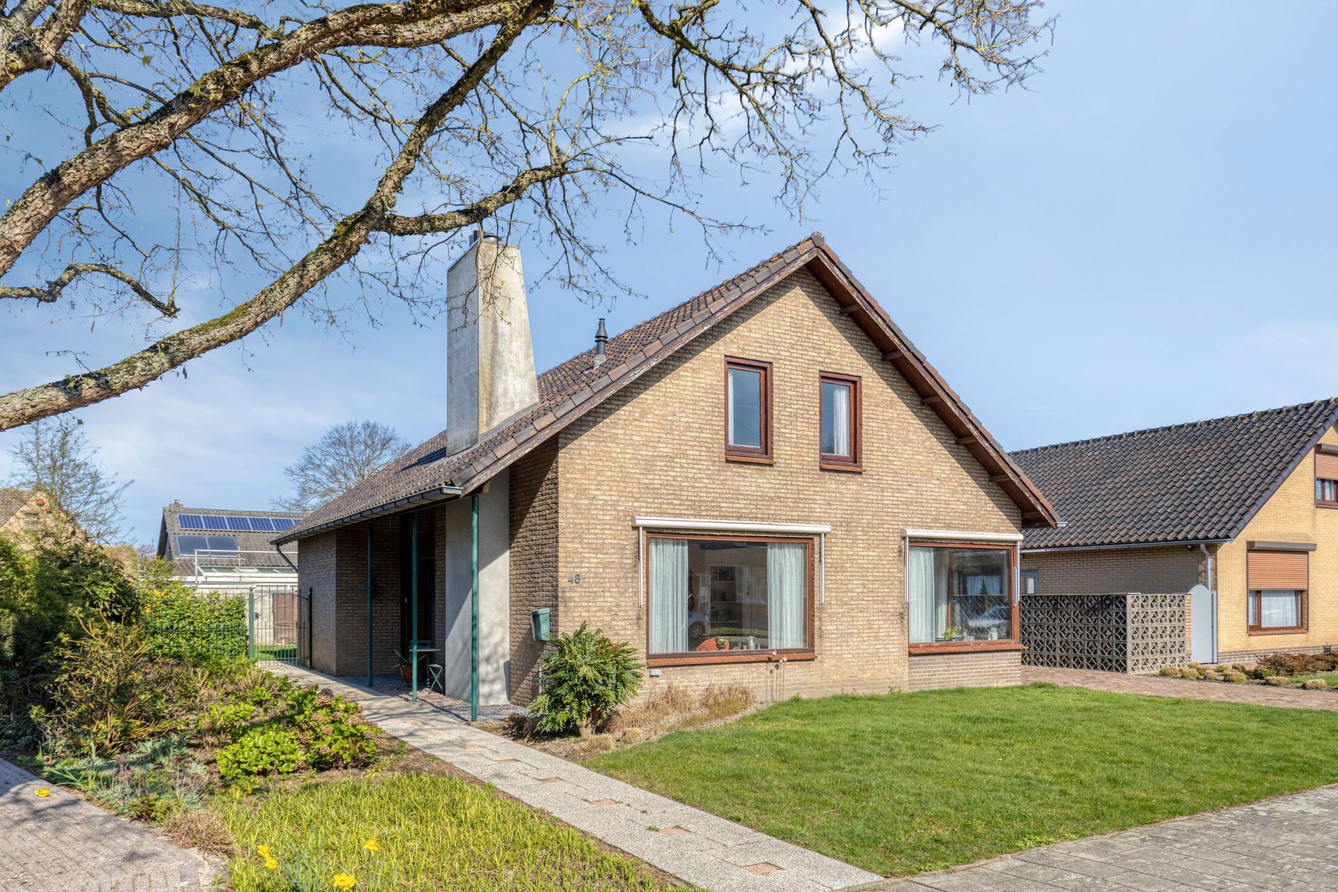 Een bakstenen huis met een steil dak, een voortuin en een geplaveid pad onder een helderblauwe hemel.