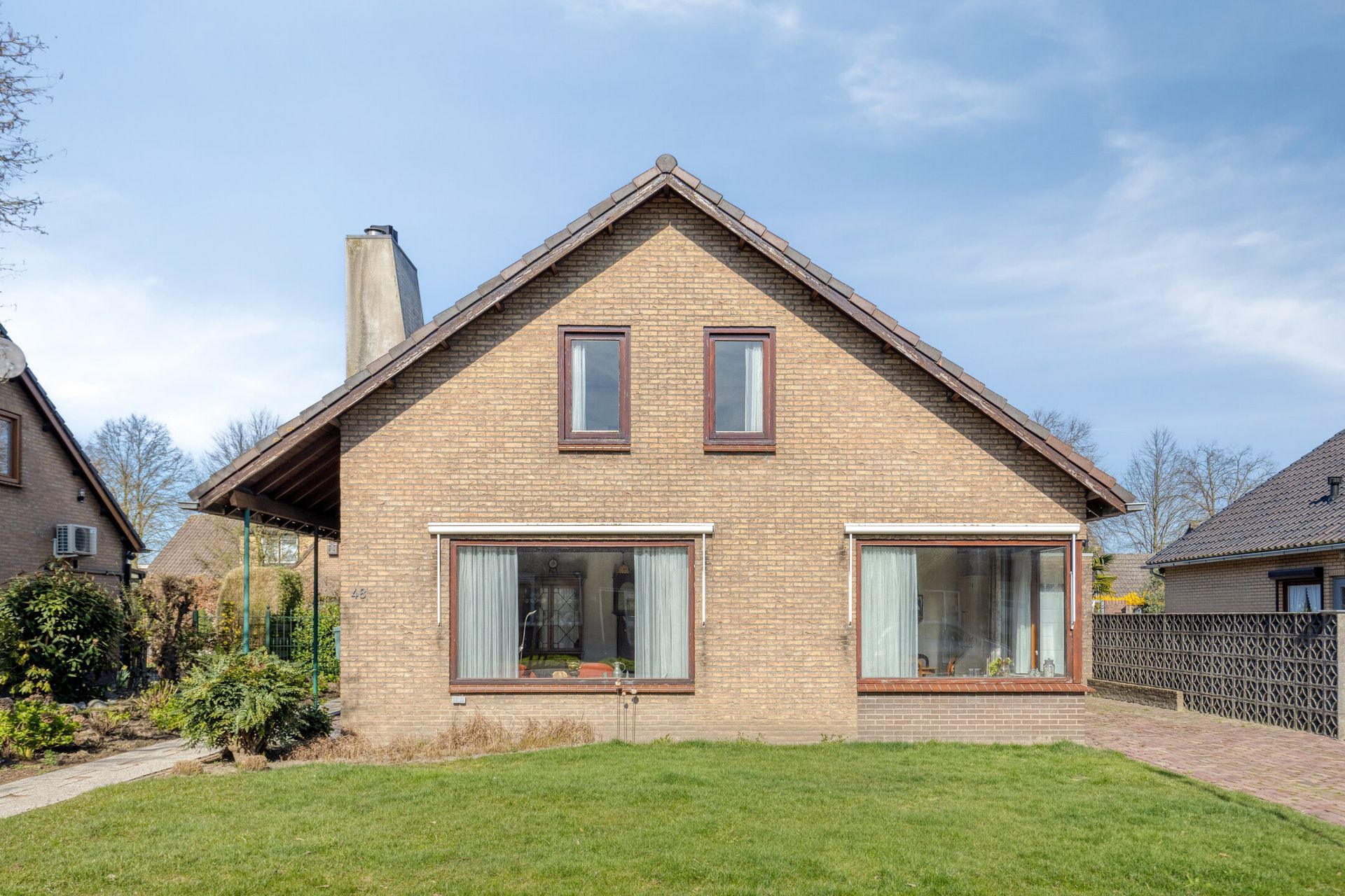 Vooraanzicht van een lichtbruin bakstenen huis met een steil dak, grote ramen en een grasveld aan de voorzijde.